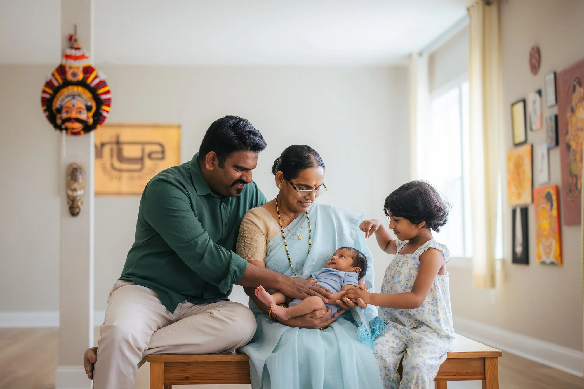 Father, grandmother in pale blue saree, and older daughter gather on a wooden bench around the newborn baby — Kathakali mask and Nritya dance studio sign visible on the wall behind them — in-home newborn session in Apex, NC.