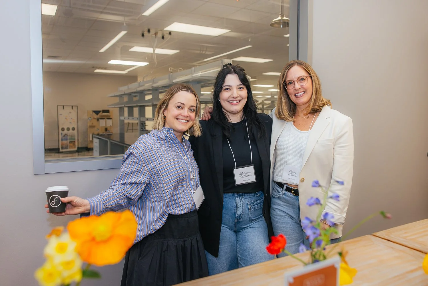 Three women smiling with name badges at a corporate event at Longfellow Triangle LIFE in Raleigh NC, colorful spring flowers in foreground