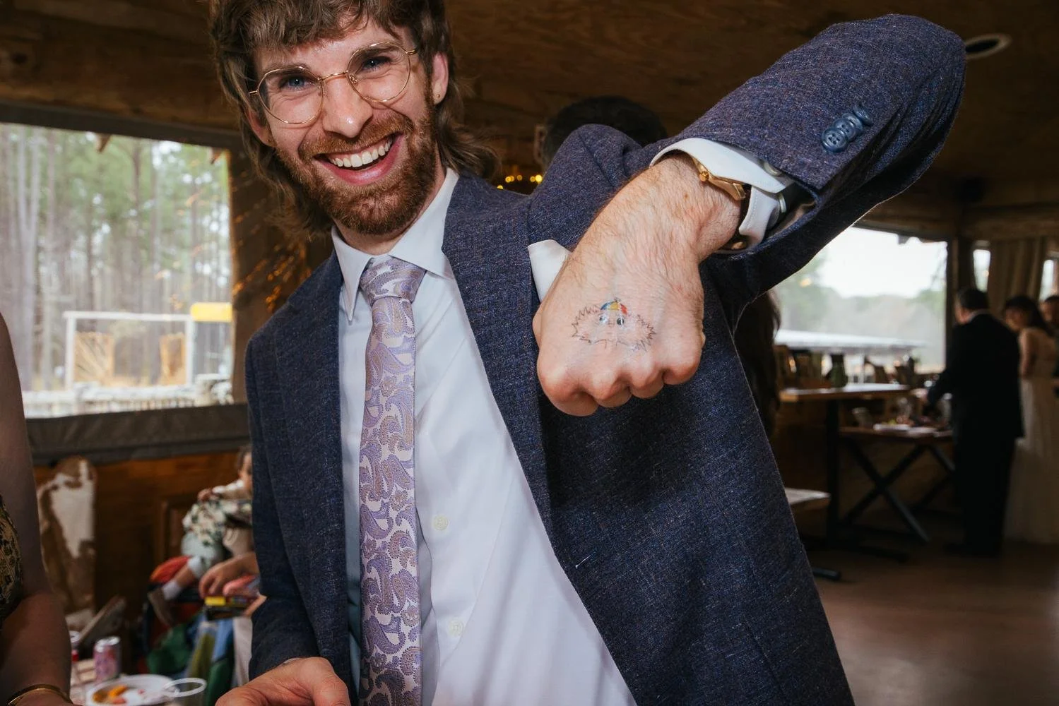 Wedding guest in a blue suit laughing and showing off a small hand tattoo during the reception at Honeysuckle Tea House Chapel Hill NC