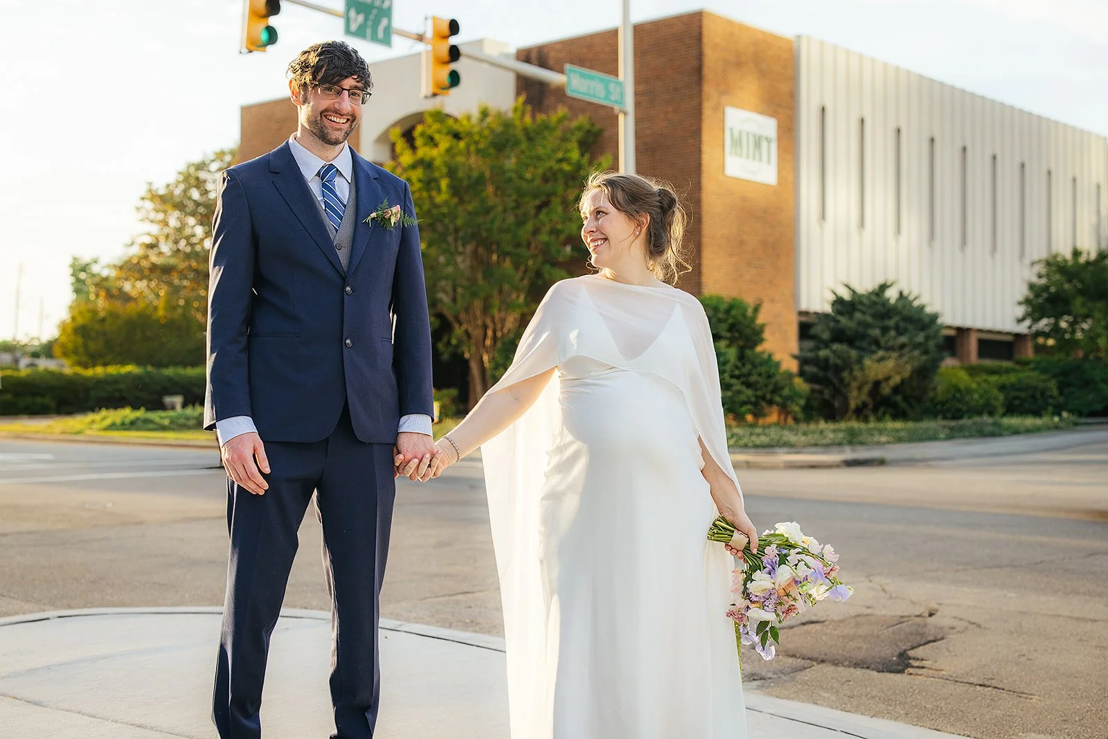 Bride and groom hold hands at a downtown Durham NC intersection at golden hour, smiling at each other, with the MINT building glowing in warm evening light behind them