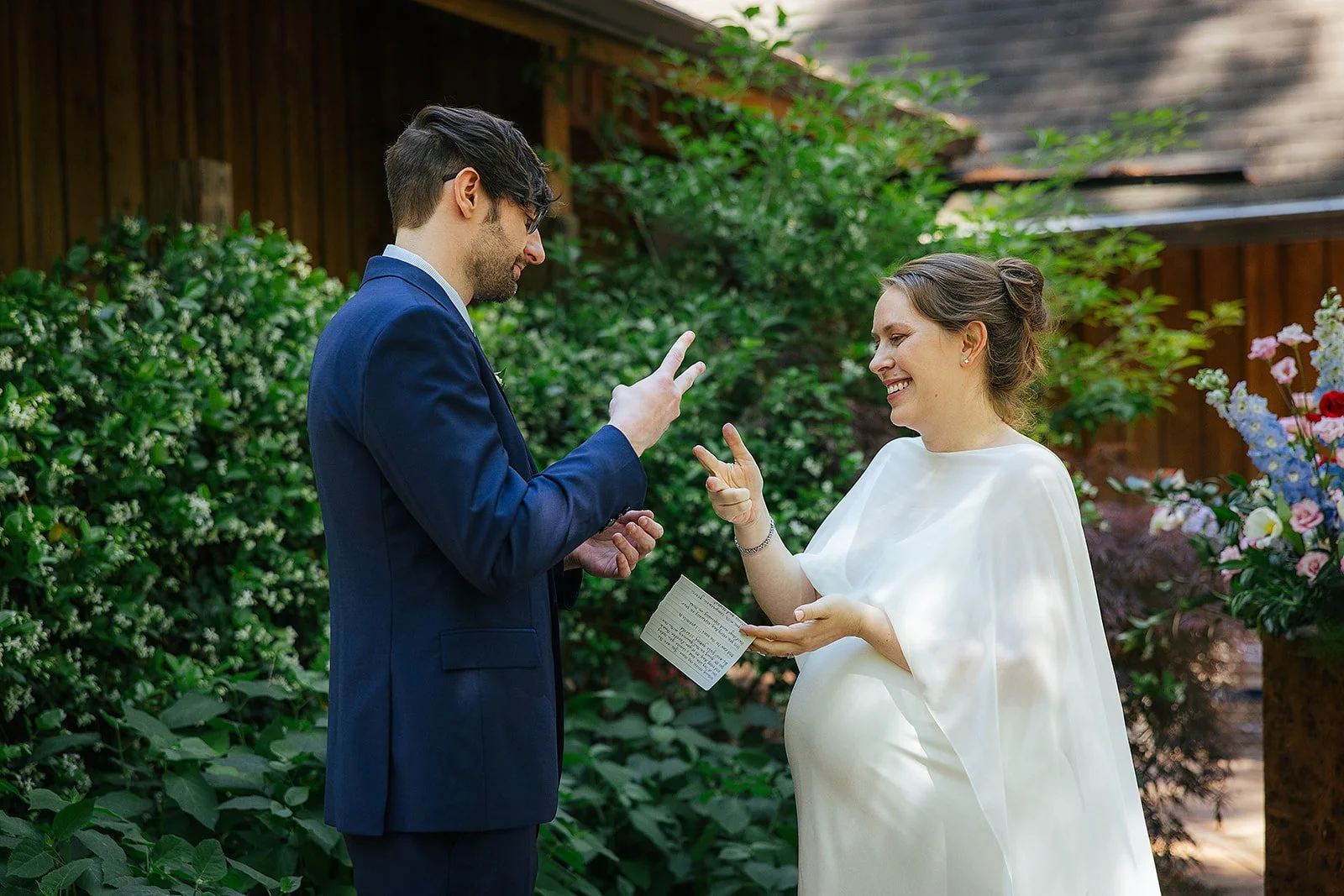 Pregnant bride throws her head back in laughter as her groom reads handwritten vows during an intimate outdoor ceremony surrounded by greenery, Durham NC