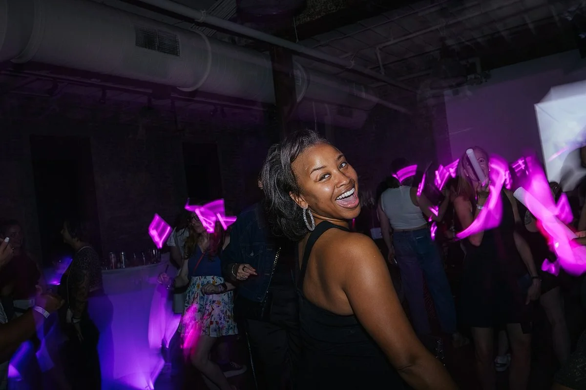 Woman glancing back over her shoulder at the camera with a full laugh, pink glow sticks lighting the crowd behind her at the Dance Her Raleigh event