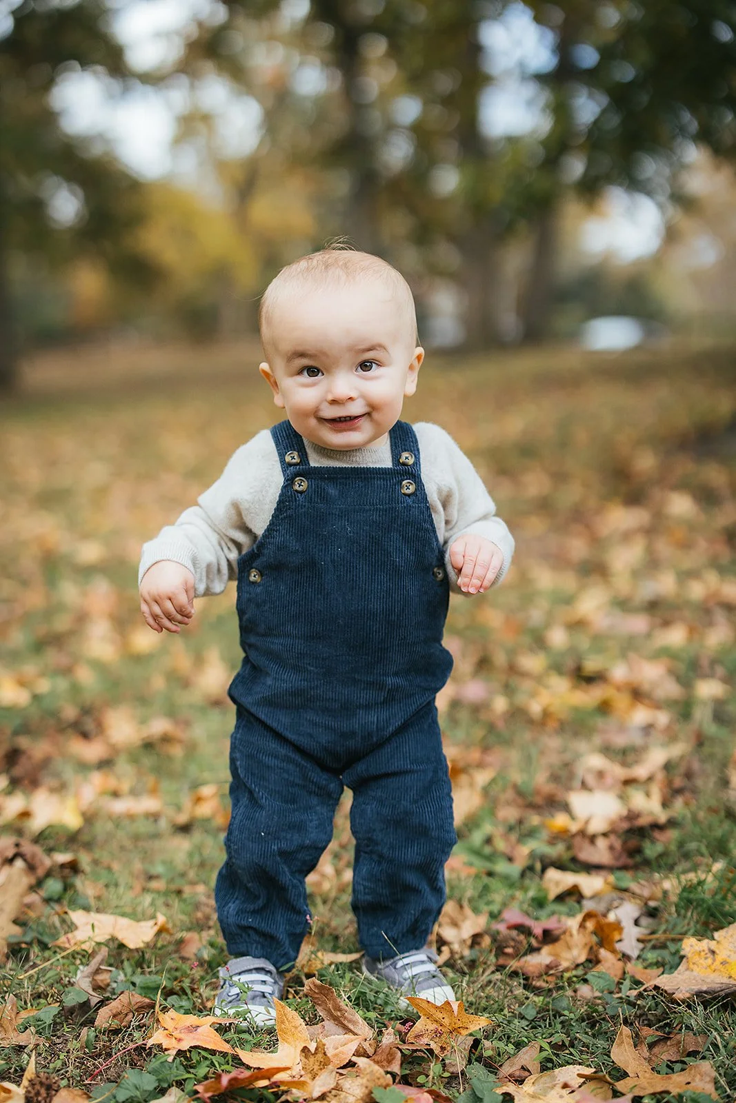 One - year - old baby boy in navy corduroy overalls standing in fall leaves at Duke Park during his first birthday session in Durham NC