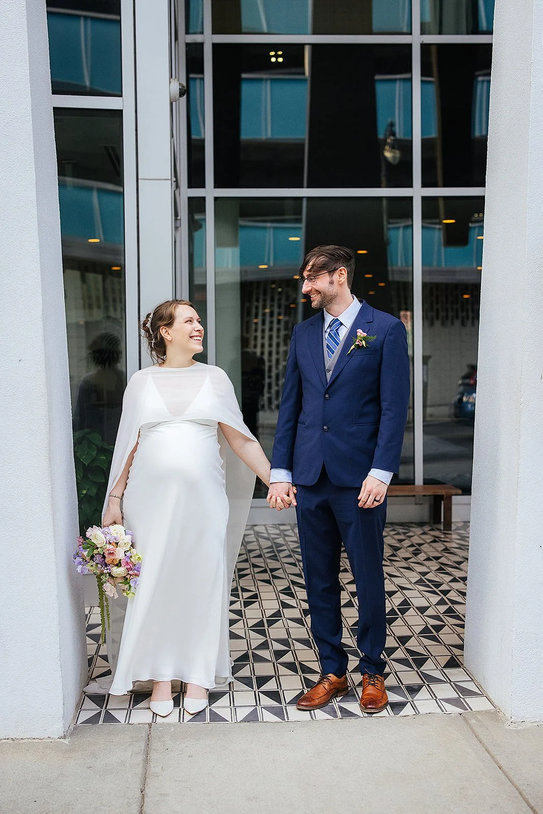 Bride and groom hold hands and smile at each other in the entrance of a downtown Durham NC building with geometric tile flooring and glass windows behind them
