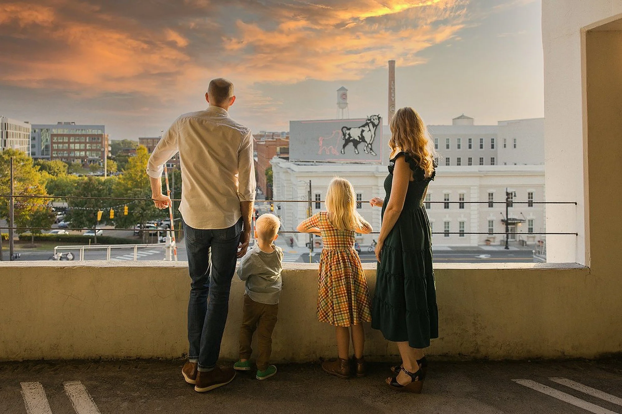Family of four in summer outfits looks out over the Durham NC skyline during a family photoshoot