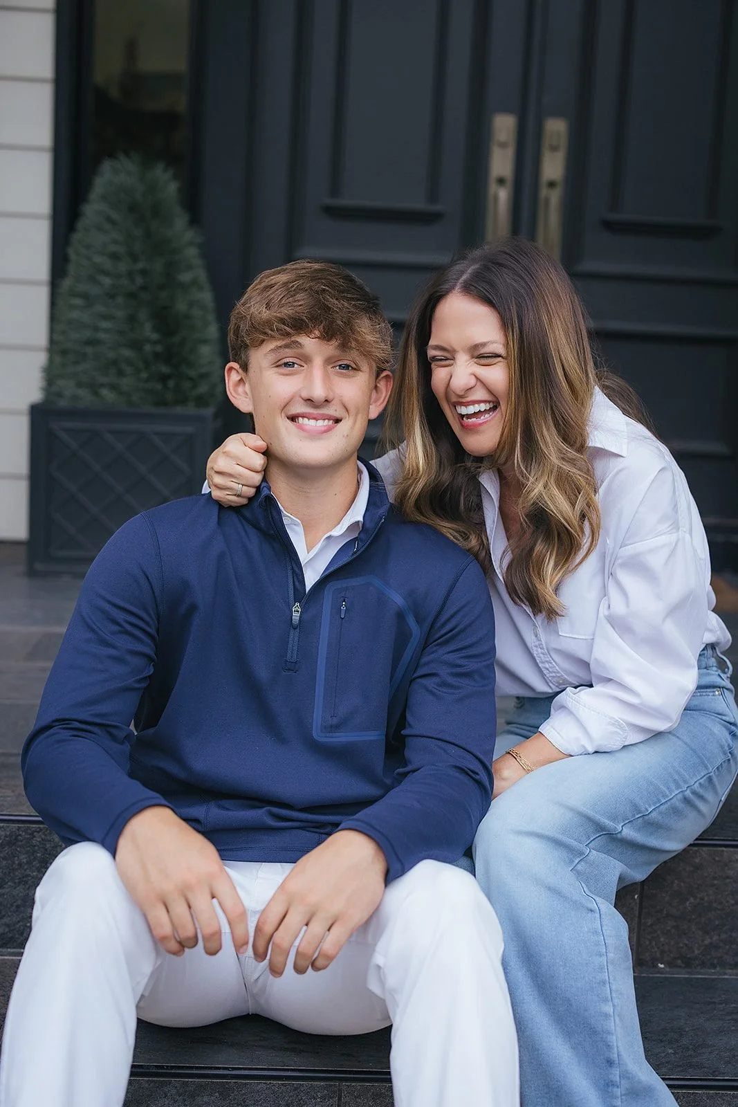 A mother throws her head back laughing with her arm around her teenage son on the front steps of their downtown Raleigh, NC home — a genuinely joyful moment between the two.