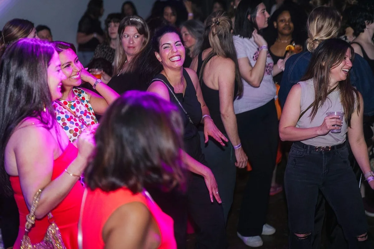 Large crowd of women smiling and dancing together under pink lights at the Dance Her event in Raleigh, NC