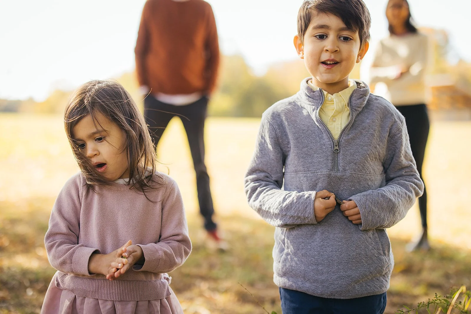 Two young siblings standing together in a bright open sunny field at Blackwood Farm Park, parents softly blurred in the background, Triangle NC family photographer