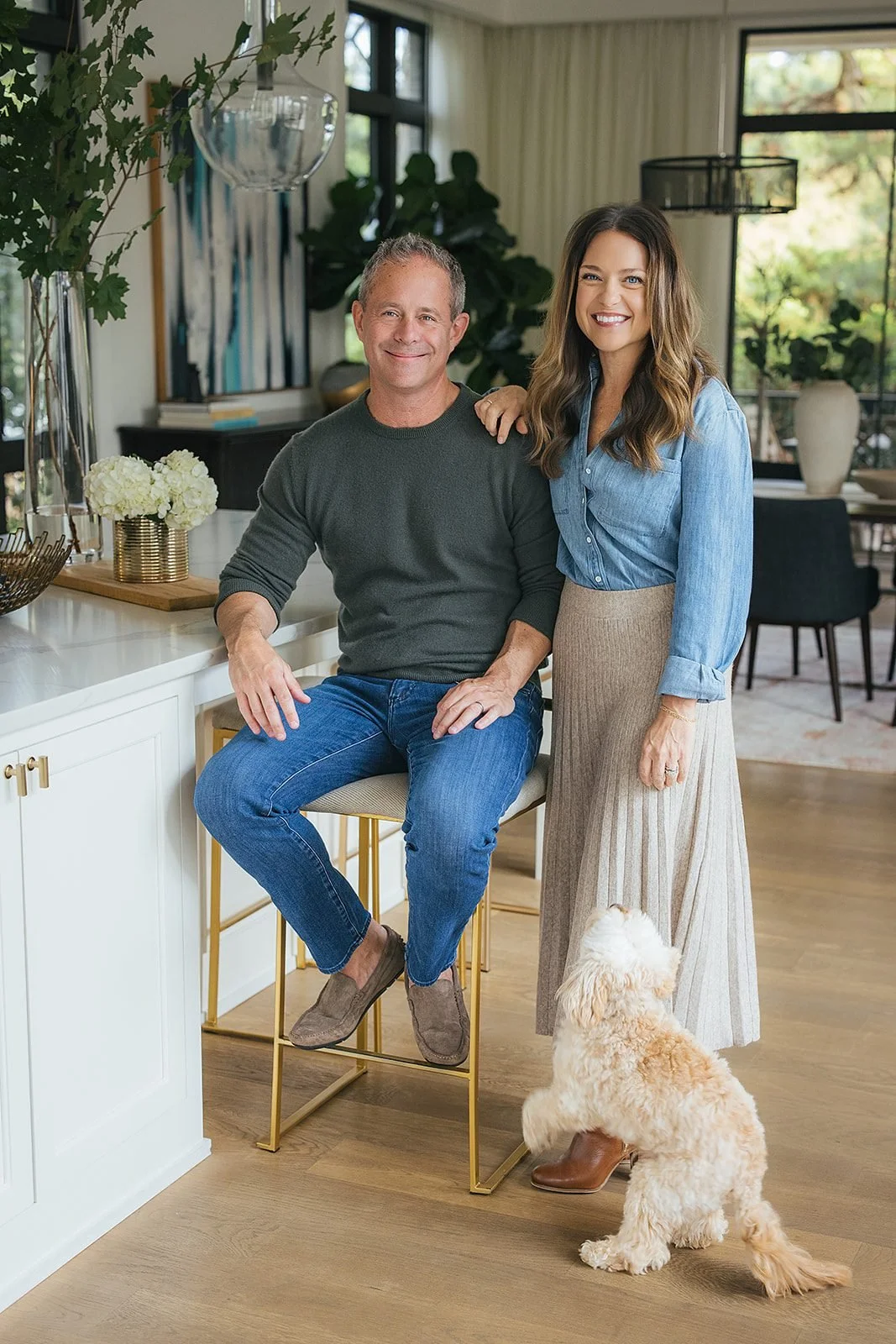 A couple stands together in their bright, modern kitchen, their goldendoodle at their feet, smiling during their in-home family session in downtown Raleigh, NC.