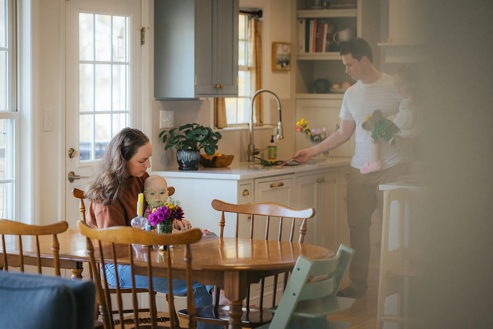 Durham family photographer captures mom with baby at kitchen table while dad cooks in the background during an in-home lifestyle session