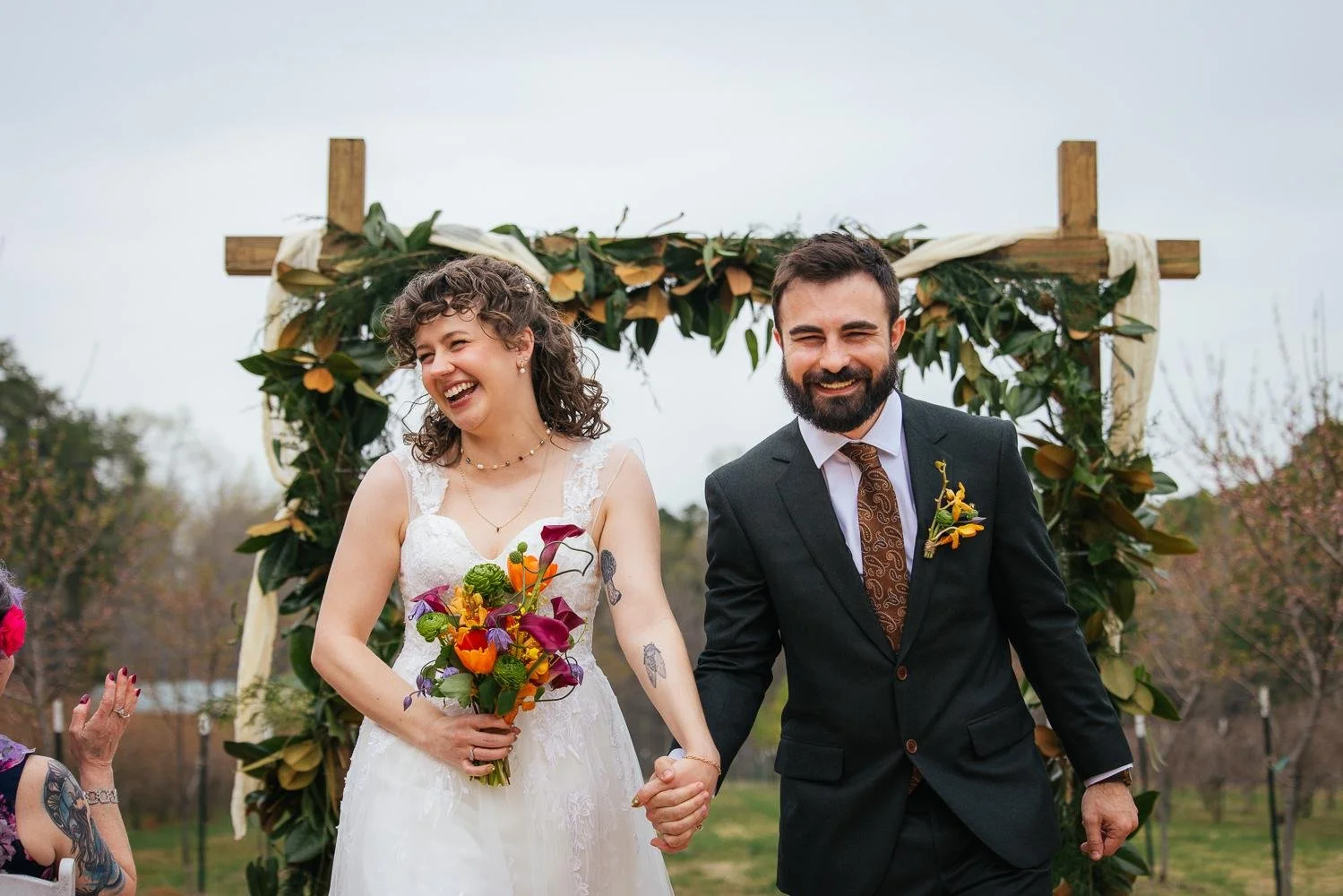 Bride and groom just married — walking recessional laughing and beaming under the magnolia and greenery floral arch at Honeysuckle Tea House outdoor ceremony Chapel Hill NC