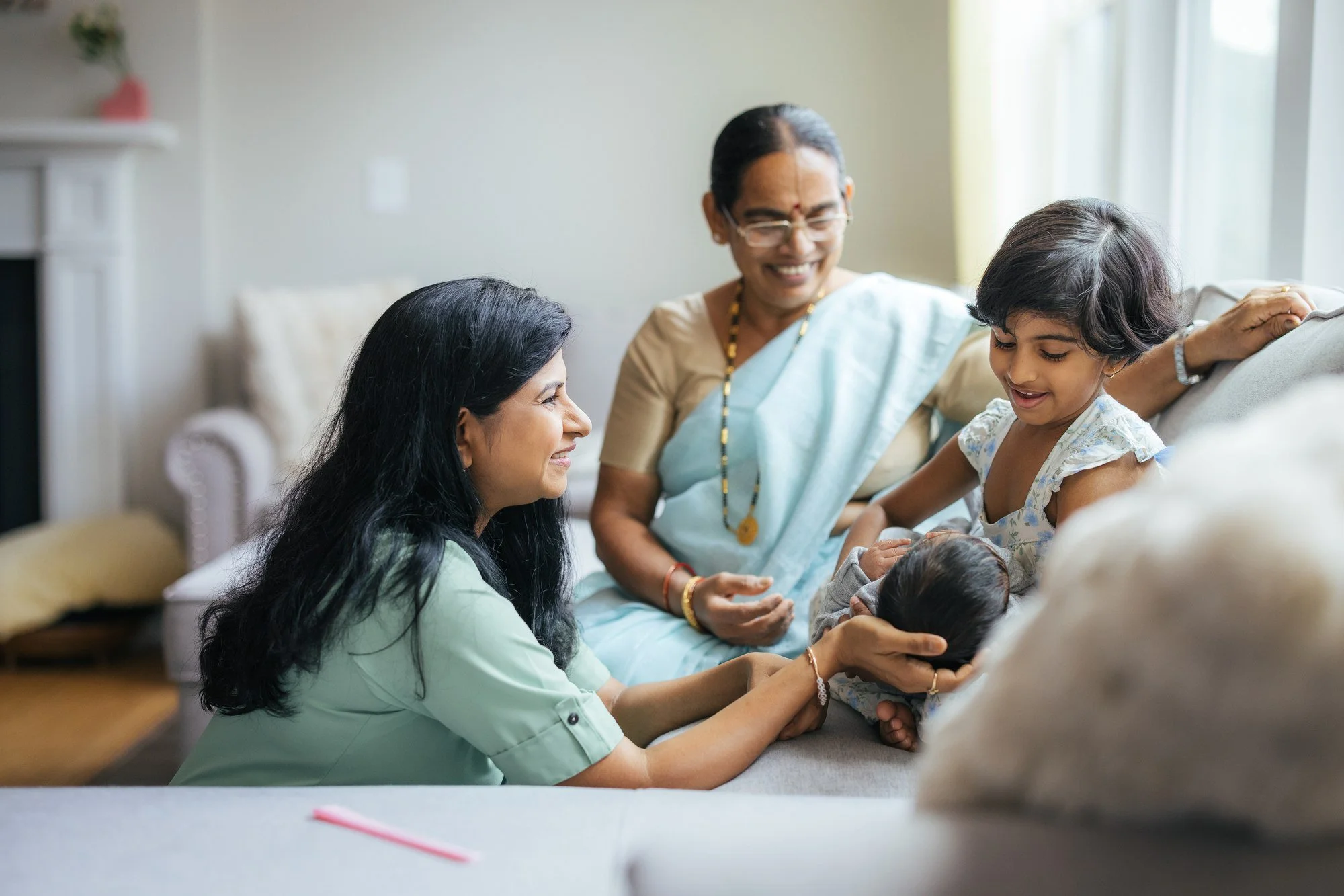 Mother in sage green, grandmother in pale blue saree, and older daughter sit together on the sofa with the newborn baby — a three-generations portrait from an in-home family session in Apex, NC.
