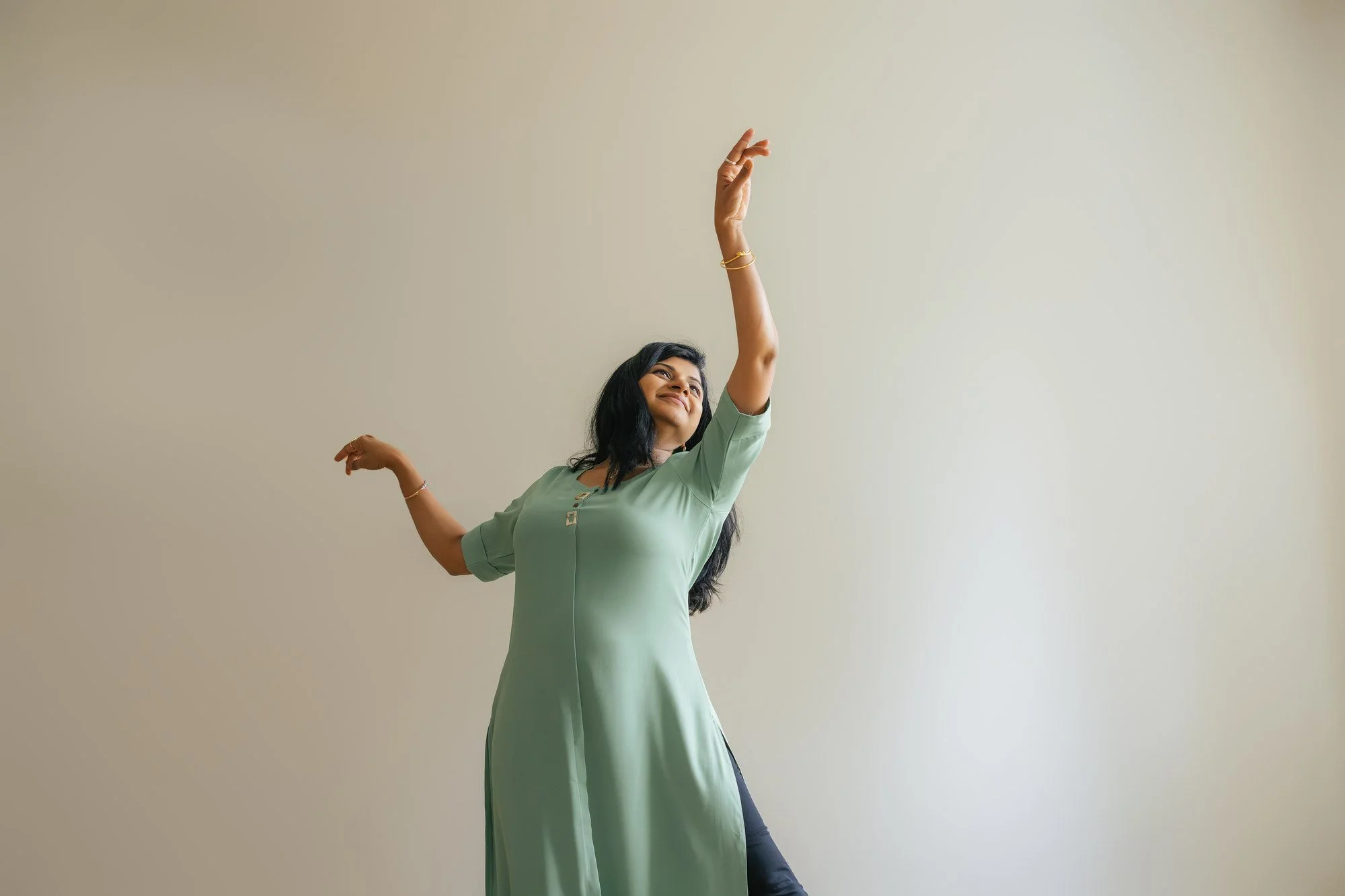 Woman in a sage green kurta stands against a soft cream wall with one arm raised gracefully overhead and the other curved at her side, smiling upward — solo portrait of a dance instructor at her in-home family session in Apex, NC.