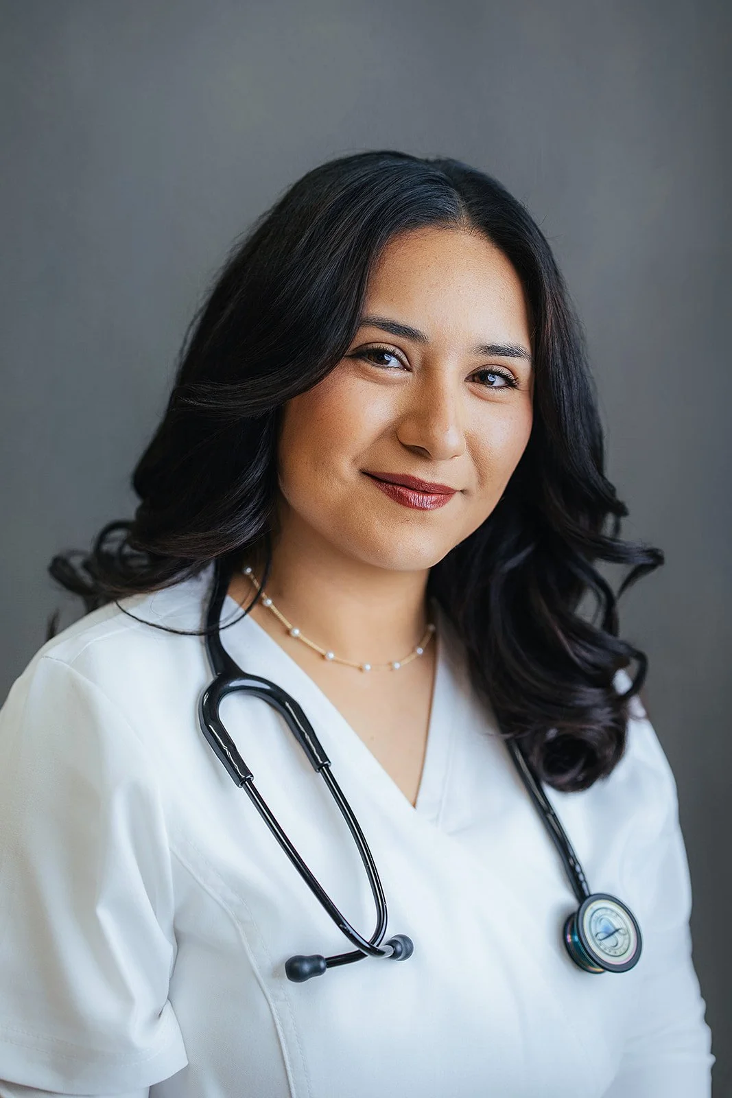 Durham Tech nursing student with long wavy dark hair and pearl necklace, soft confident smile in white scrubs, professional headshot in Durham NC