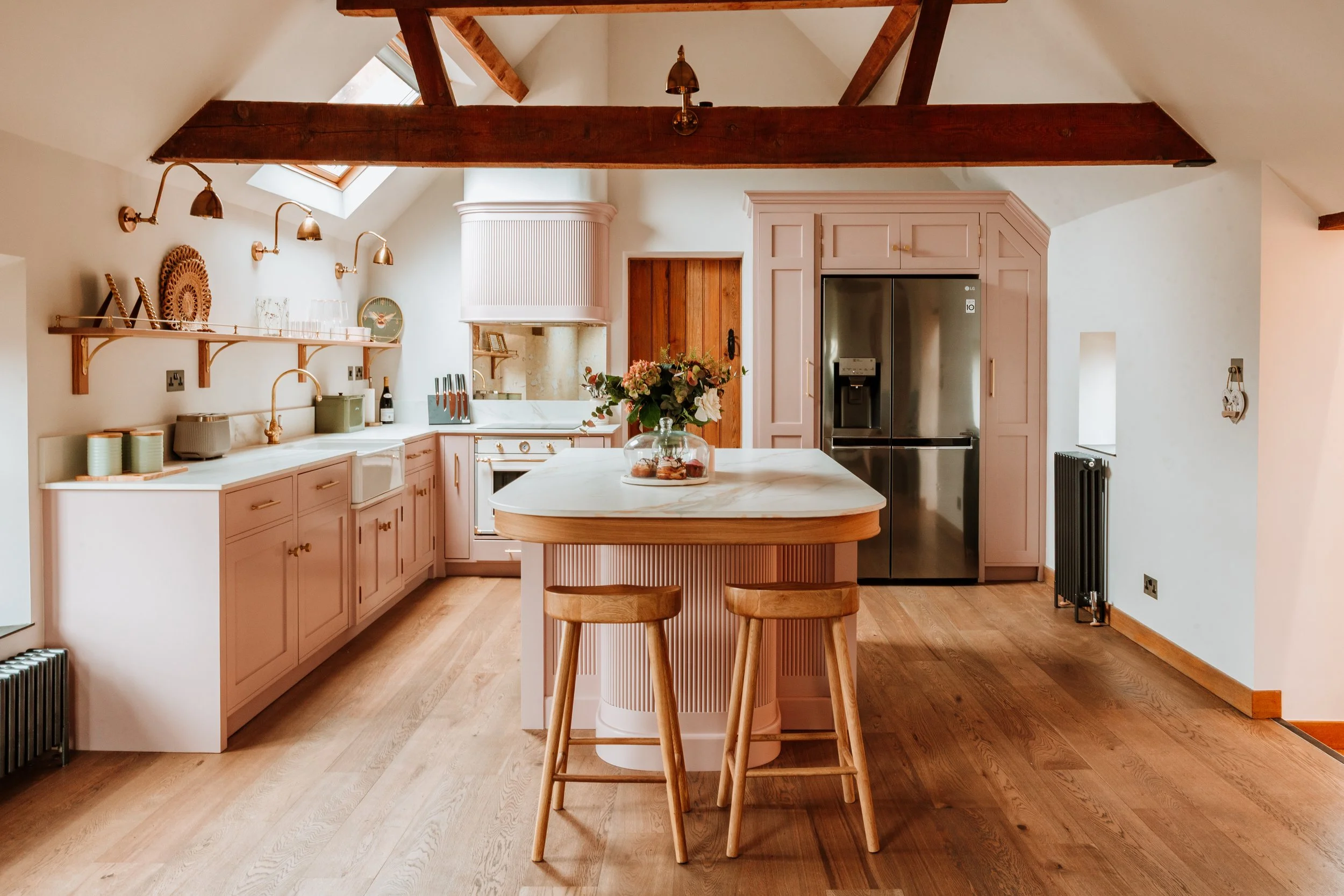 A cozy kitchen with light pink cabinets, a marble island with two wooden barstools, a stainless steel refrigerator, oven, and a vase of flowers on the island. The room features wooden beams and hardwood flooring. Interiors by Lucy Rebecca Photography