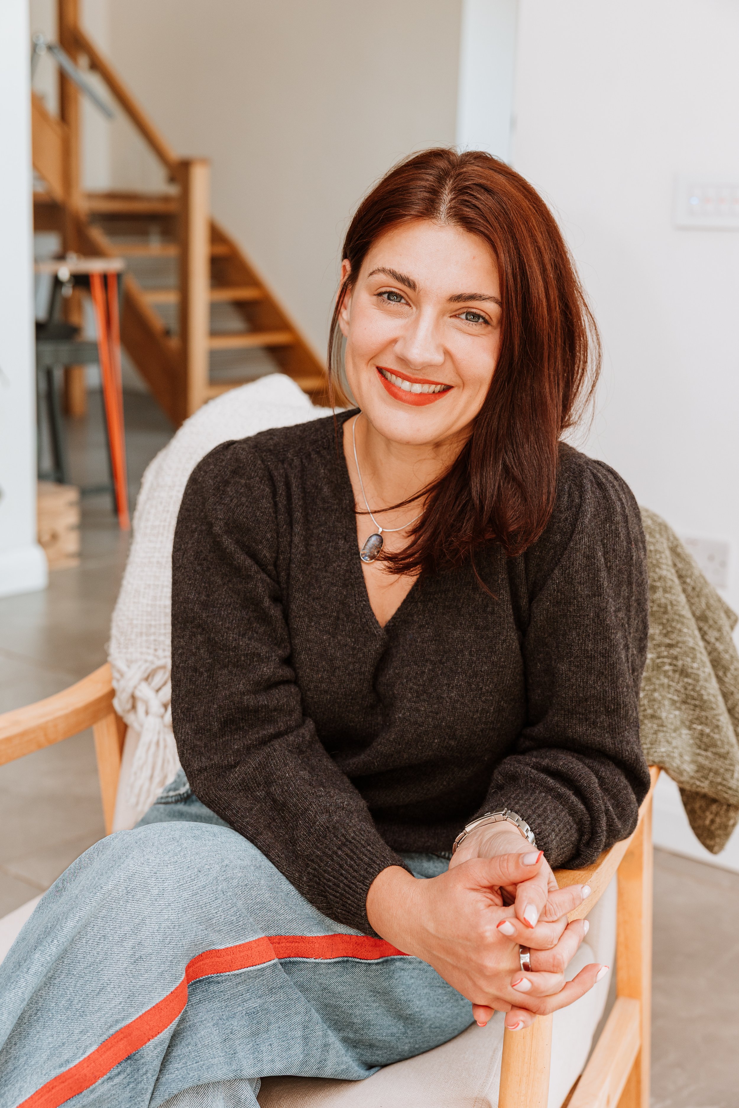 A woman with auburn hair and red lipstick smiling, sitting in a modern home interior. Personal Branding by Lucy Rebecca Photography.