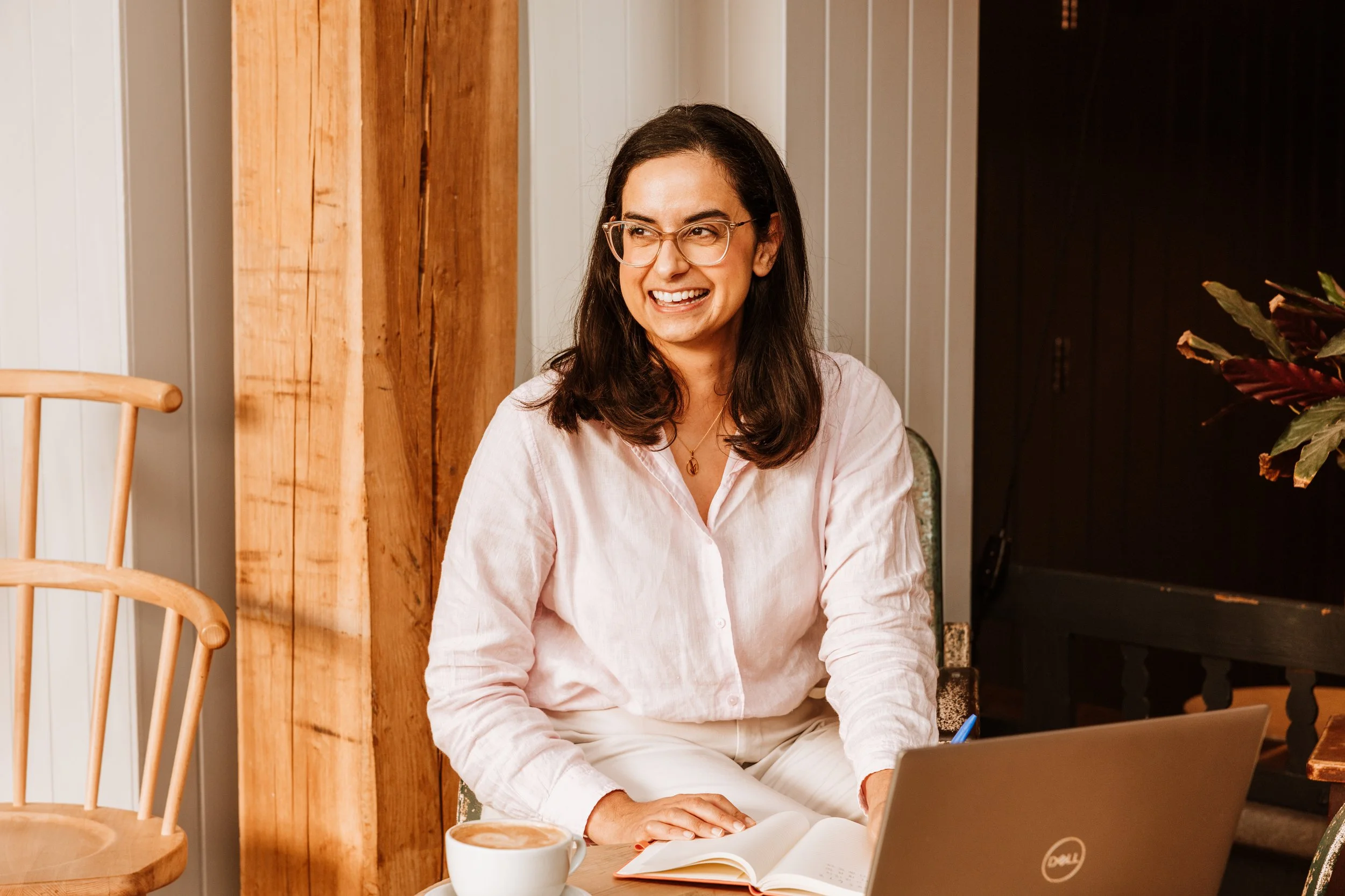 Woman with glasses smiling, sitting at a table with a laptop, open notebook, and a coffee cup in a cozy, wooden interior. Personal Branding by Lucy Rebecca Photography.