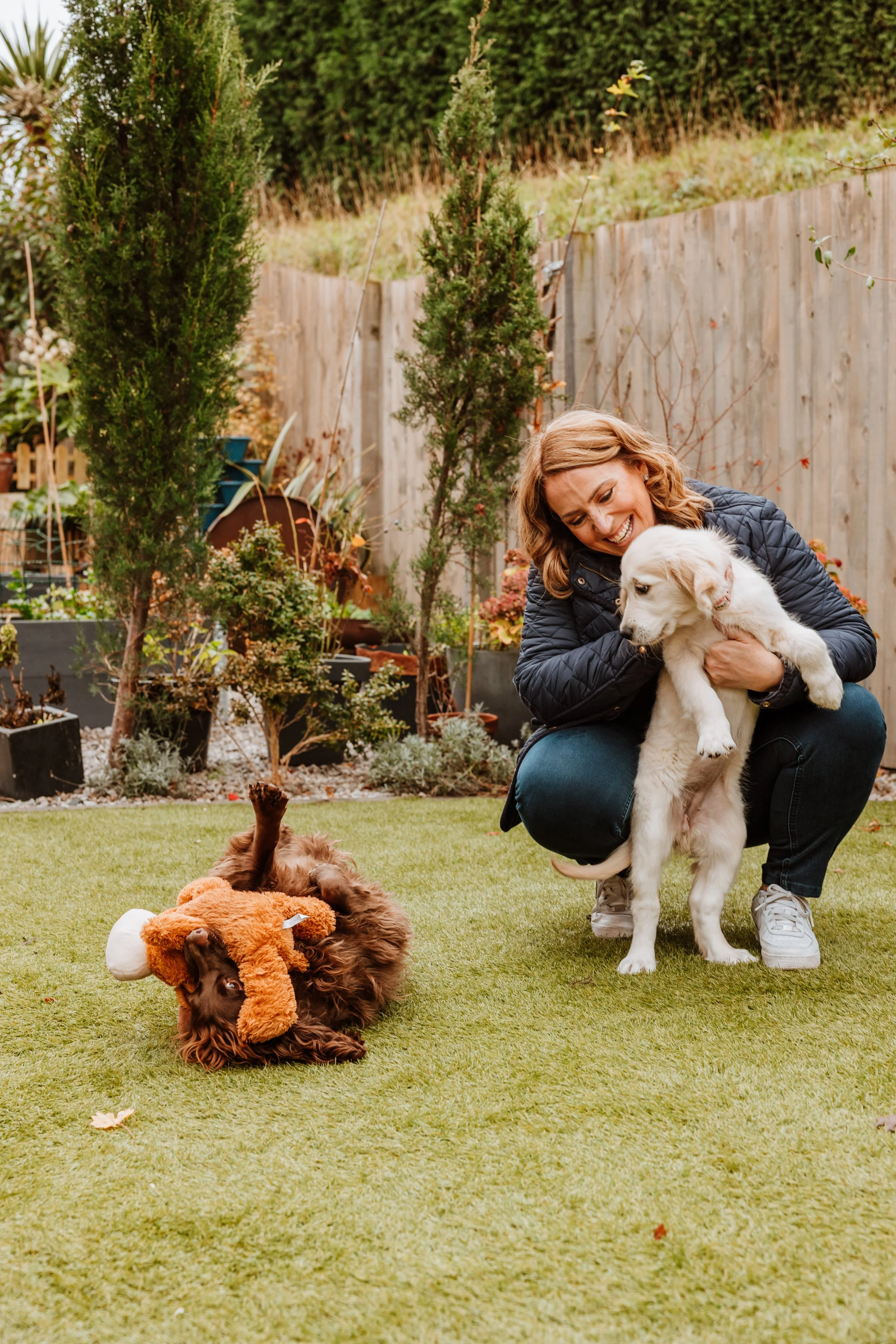 A woman playing with two puppies in a backyard with grass, trees, and a wooden fence. Personal Branding by Lucy Rebecca Photography.