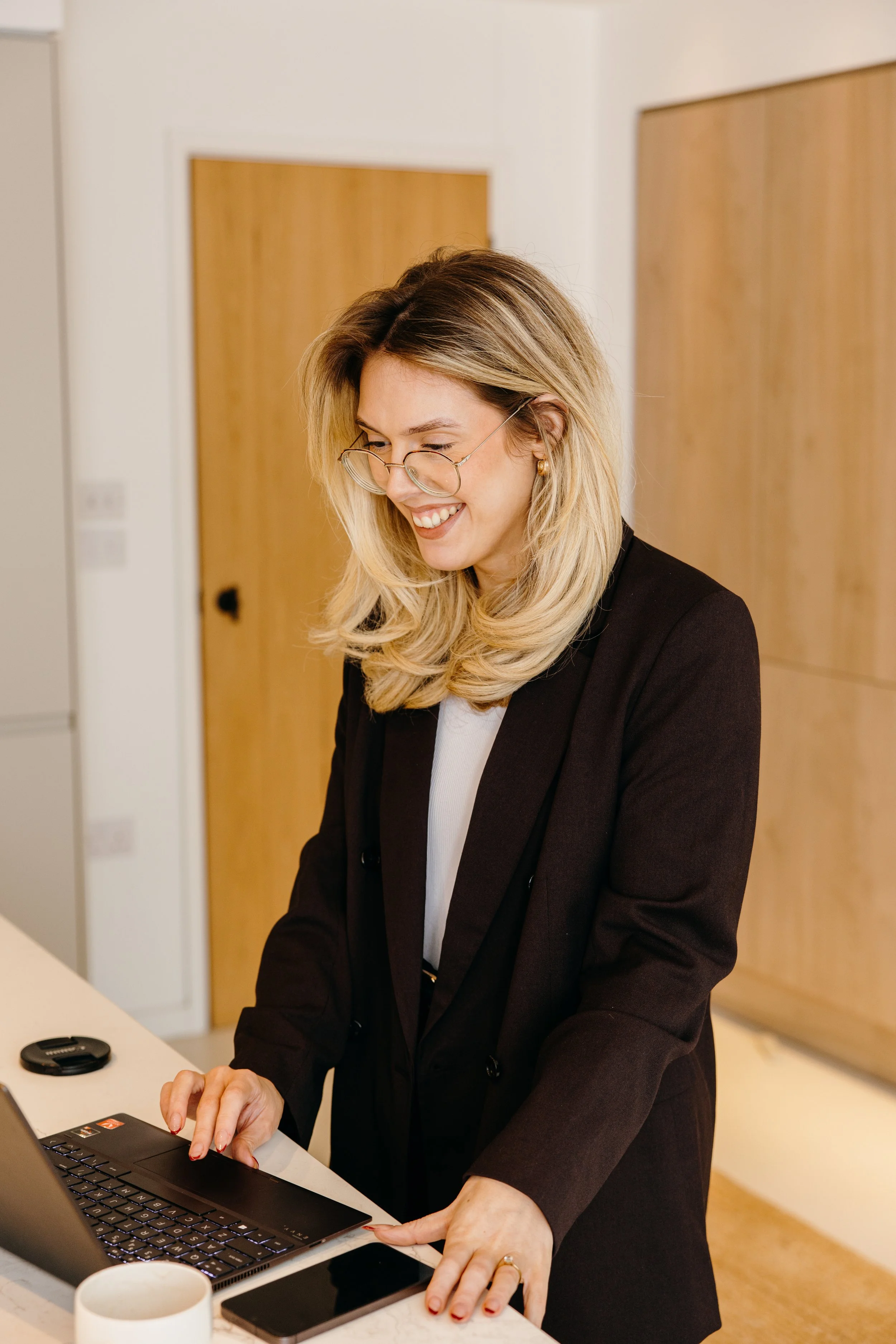 A woman with blonde hair and glasses smiling and working on a laptop in a modern kitchen. Personal Branding by Lucy Rebecca Photography.