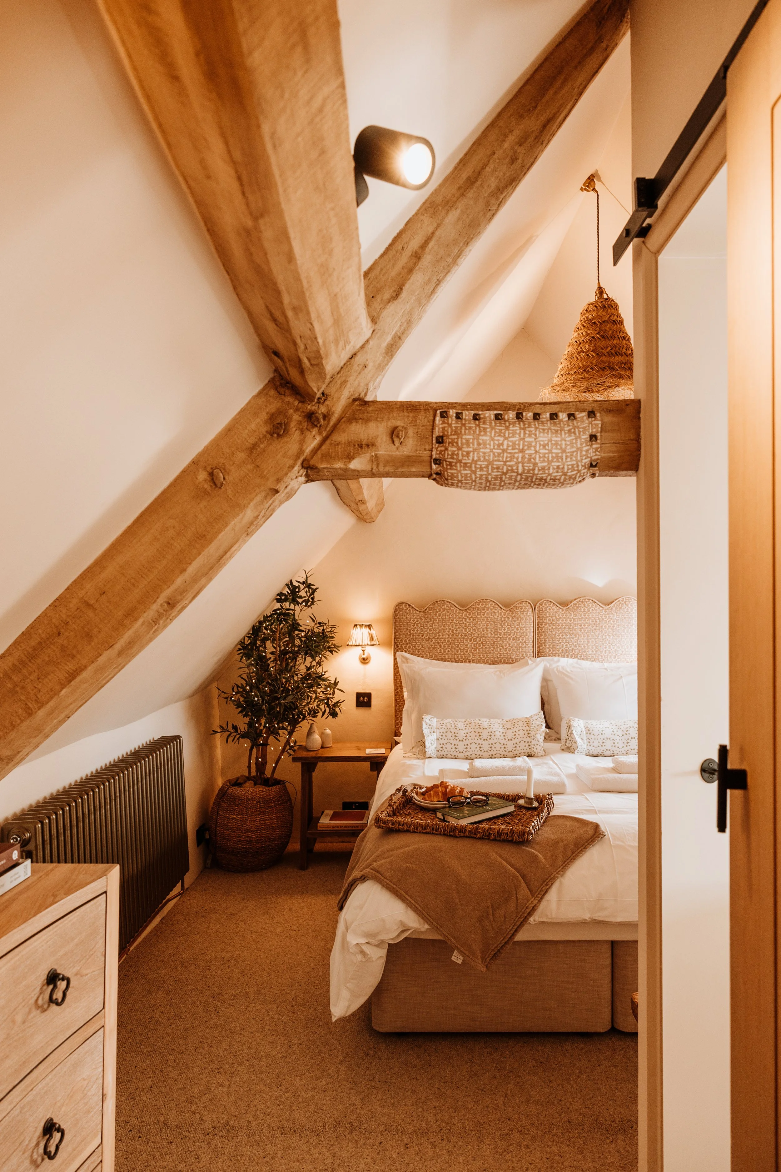 Cozy bedroom with a sloped ceiling, exposed wooden beams, and warm lighting. The bed has a padded headboard, white linens, and decorative pillows. Beside the bed is a small nightstand with a lamp, a potted plant, and a few books. A tray with reading 
