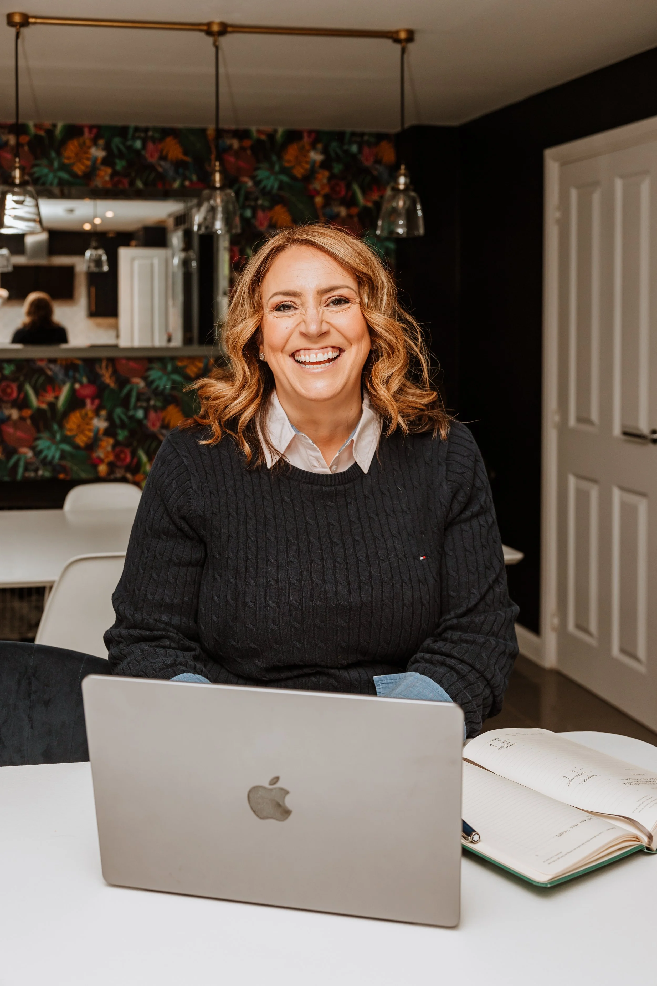 A woman with shoulder-length curly red hair smiling while sitting at a table with a silver MacBook and an open notebook in front of her, in a room with a dark floral wallpaper and hanging pendant lights. Personal Branding by Lucy Rebecca Photography.