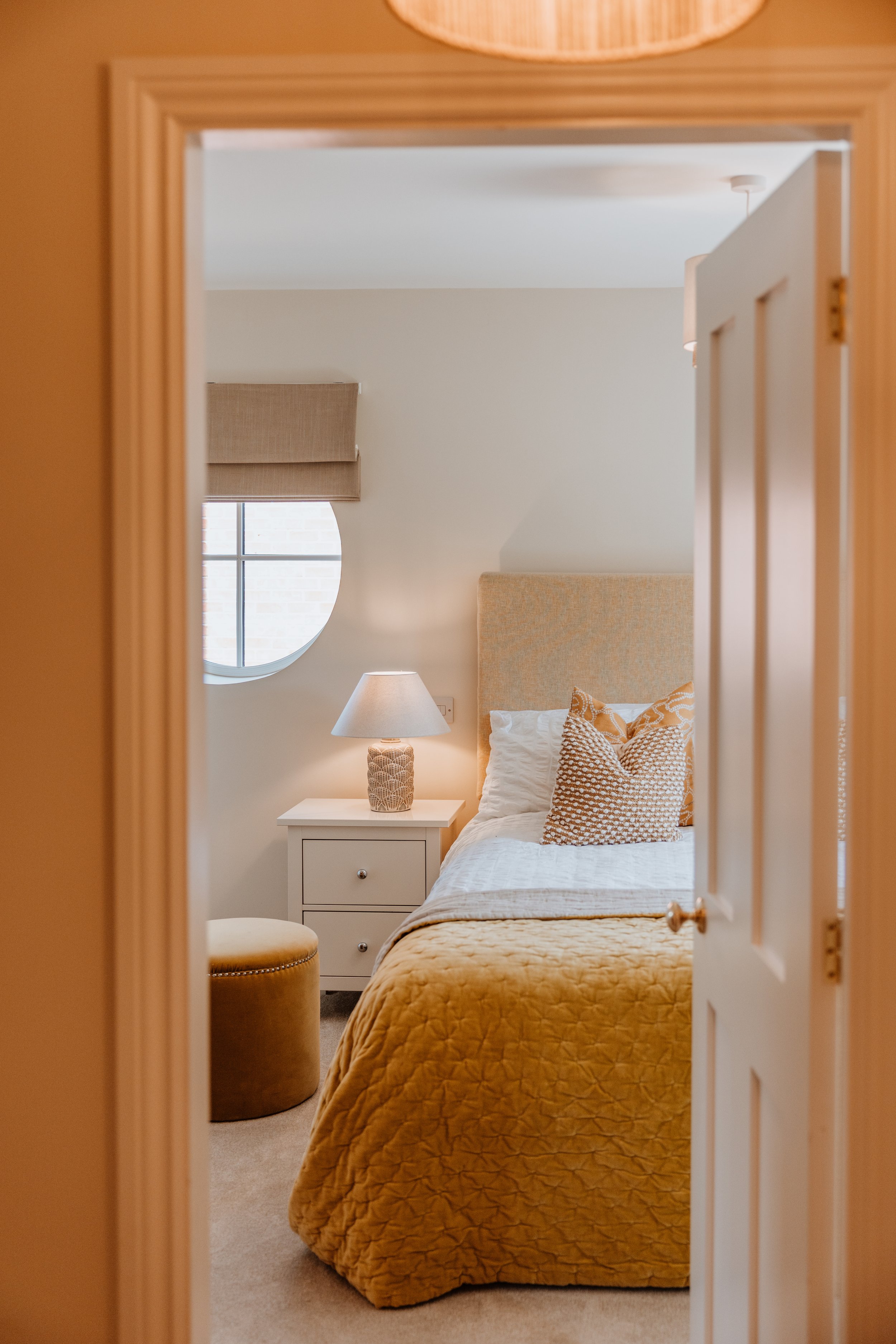 View of a cozy bedroom through an open doorway, featuring a bed with a yellow quilt, patterned pillows, a white nightstand with a lamp, a window with a round top, and a beige upholstered ottoman. Interiors by Lucy Rebecca Photography.