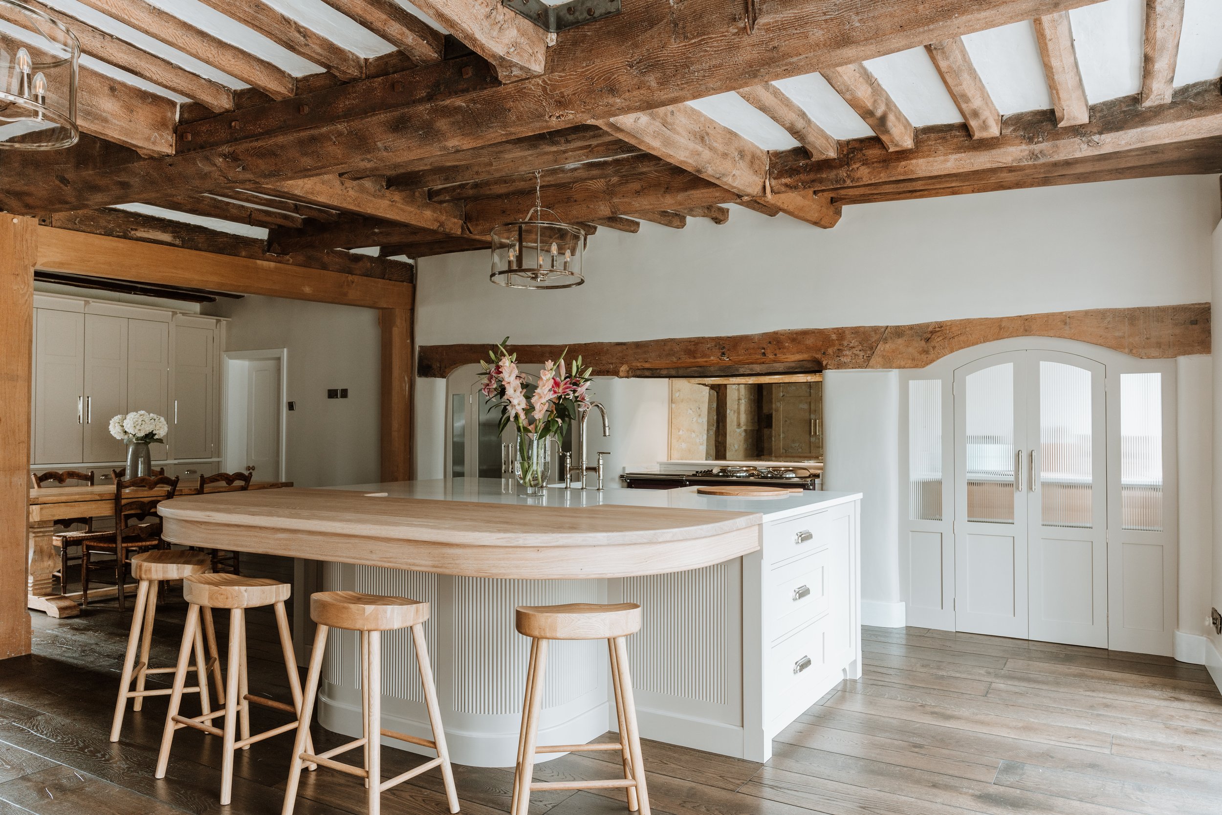 A rustic kitchen with exposed wooden beams, a white island with a wooden countertop, and wooden barstools. There are flowers in a vase on the island, and a dining area with chairs and a table in the background. Interiors by Lucy Rebecca Photography.