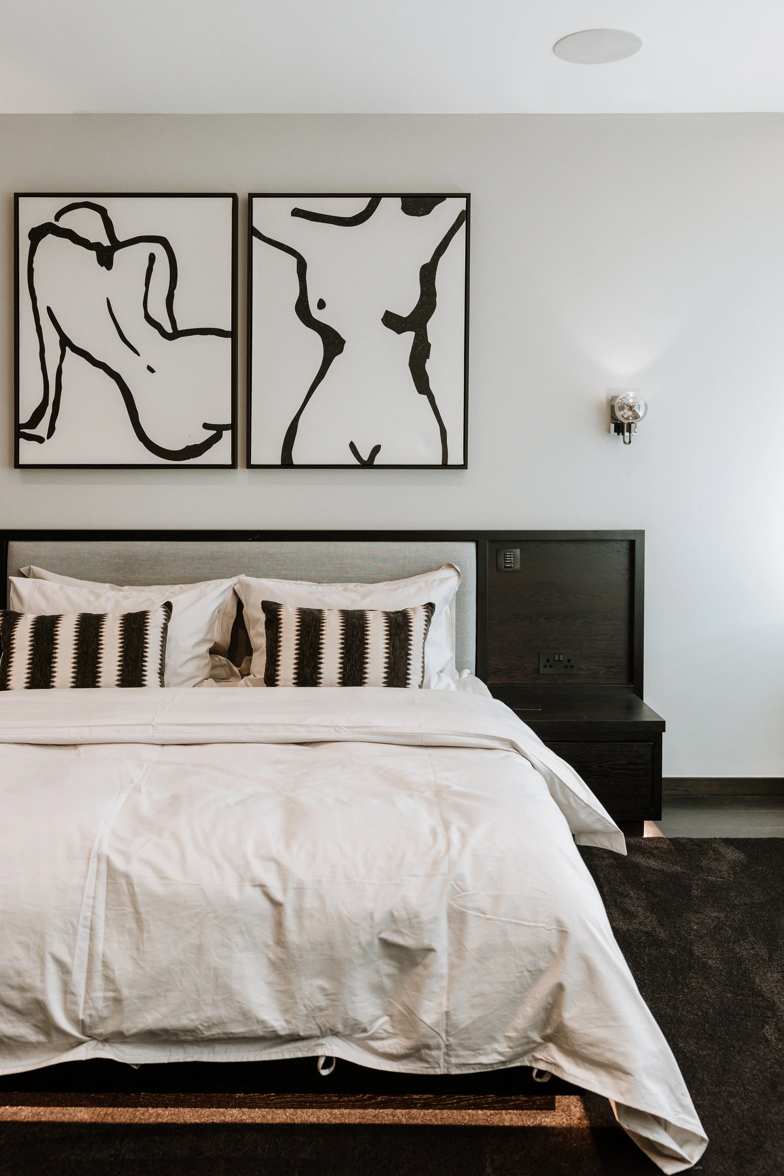 A modern bedroom with a bed, white bedding, and black-and-white striped pillows. Above the bed are two abstract black-and-white line art paintings of female torsos. There is a small wall sconce light next to the paintings. Interiors by Lucy Rebecca 