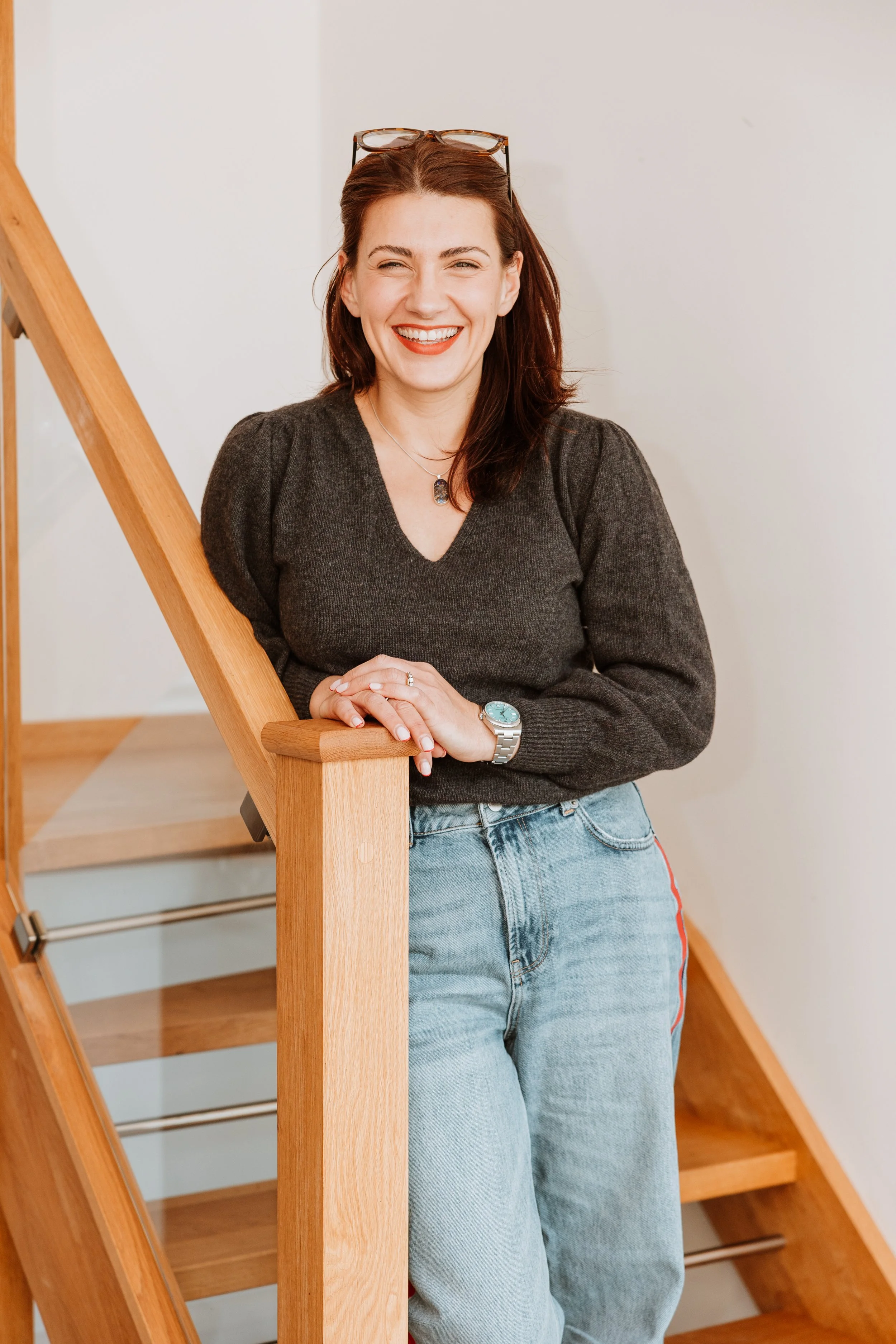 A woman with red hair and glasses resting on her head, smiling, wearing a dark sweater, watch, and pants, standing next to a wooden staircase. Personal Branding by Lucy Rebecca Photography.
