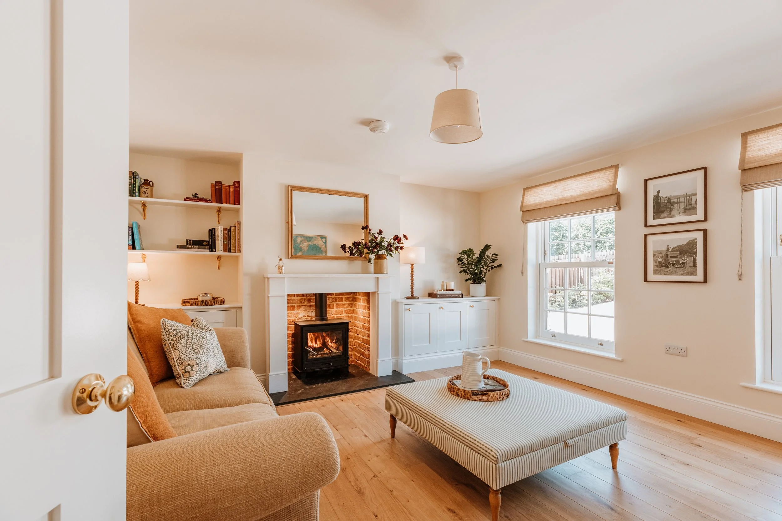Living room with a fireplace, beige sofa, striped ottoman, and windows. Interiors by Lucy Rebecca Photography.