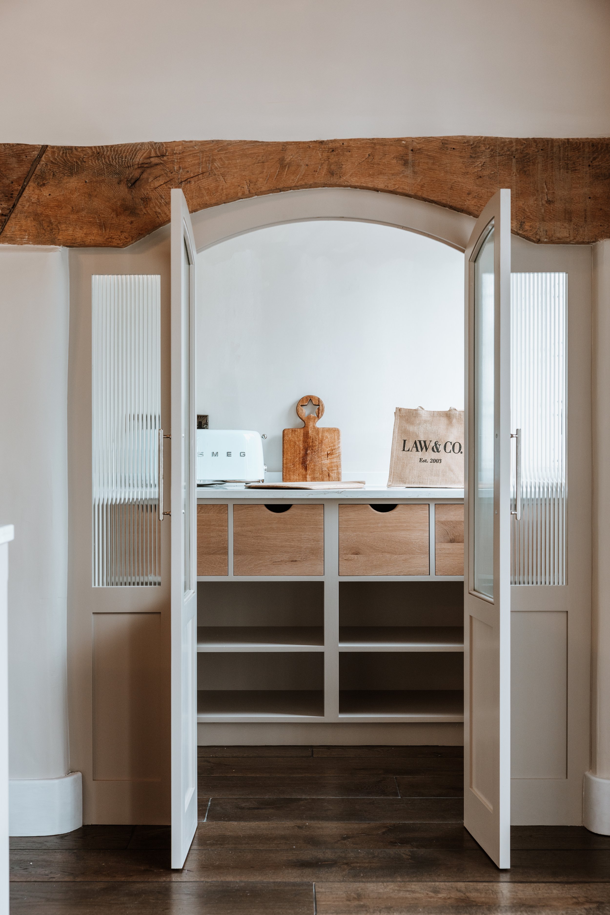 Open white double doors leading into a kitchen nook with wooden accents, a countertop, a small wooden cutting board with a star cutout, a Smeg toaster, and a paper bag labeled "LAW & CO."  Interiors by Lucy Rebecca Photography.