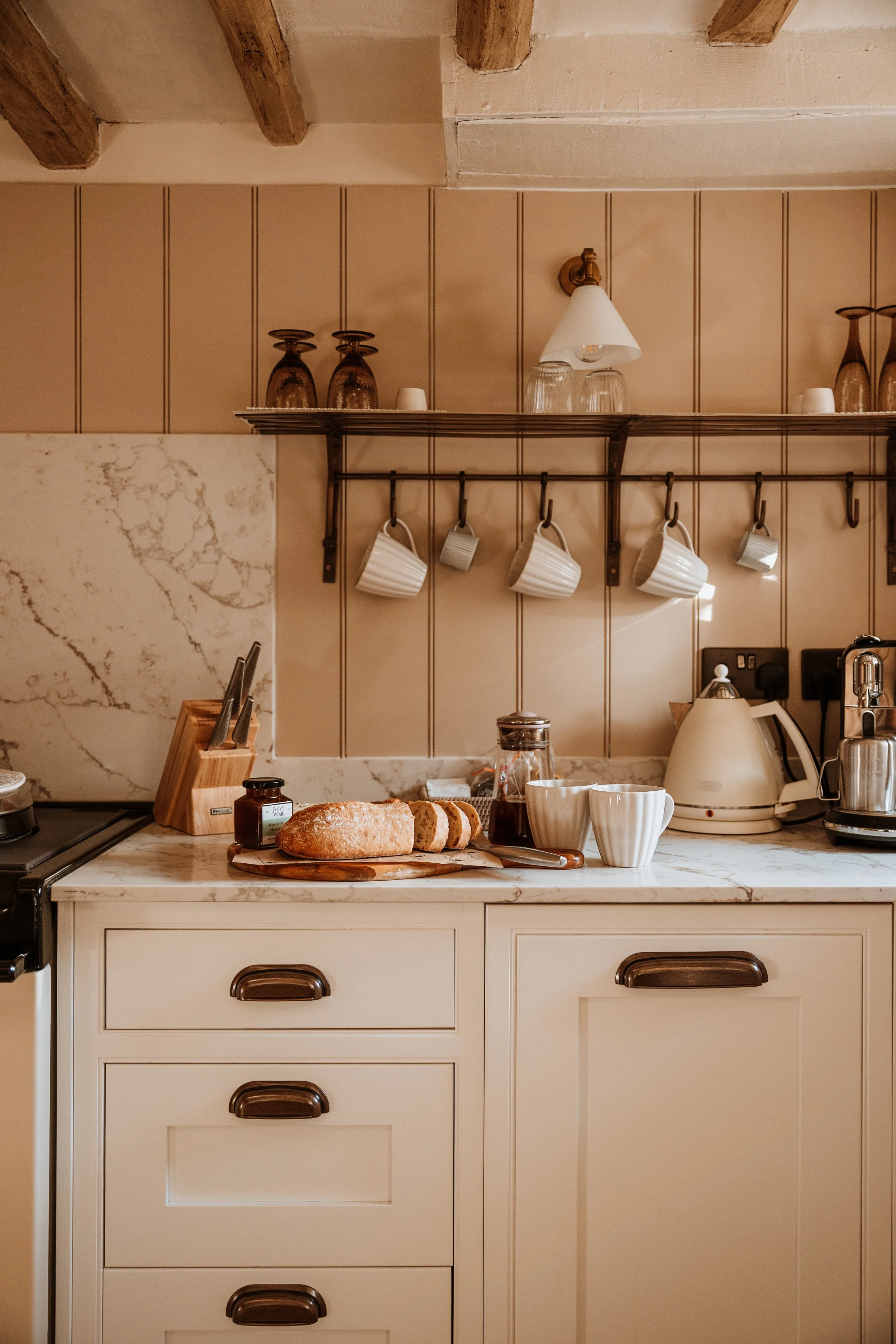 Kitchen with beige cabinetry, marble countertop, sliced bread, jars, cups, and tea kettle, with wall-mounted cups hanging from a rail and decorative glasses on a shelf. Interiors by Lucy Rebecca Photography.