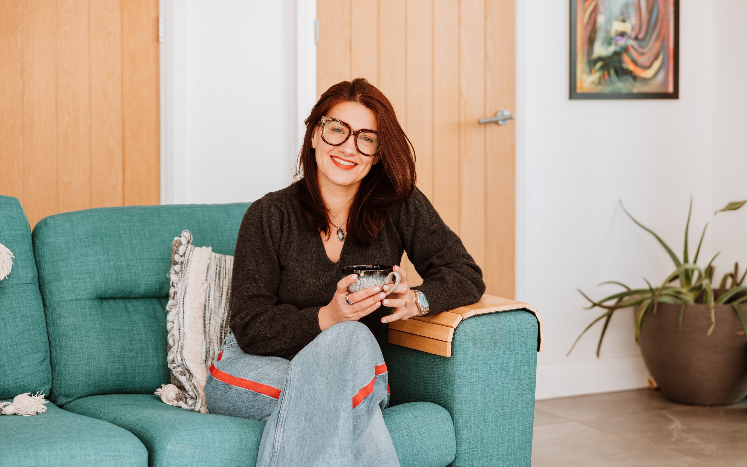 A woman with glasses and red hair sitting on a teal sofa, smiling and holding a mug, in a cozy living room with wooden panel walls and a potted plant. Personal Branding by Lucy Rebecca Photography.