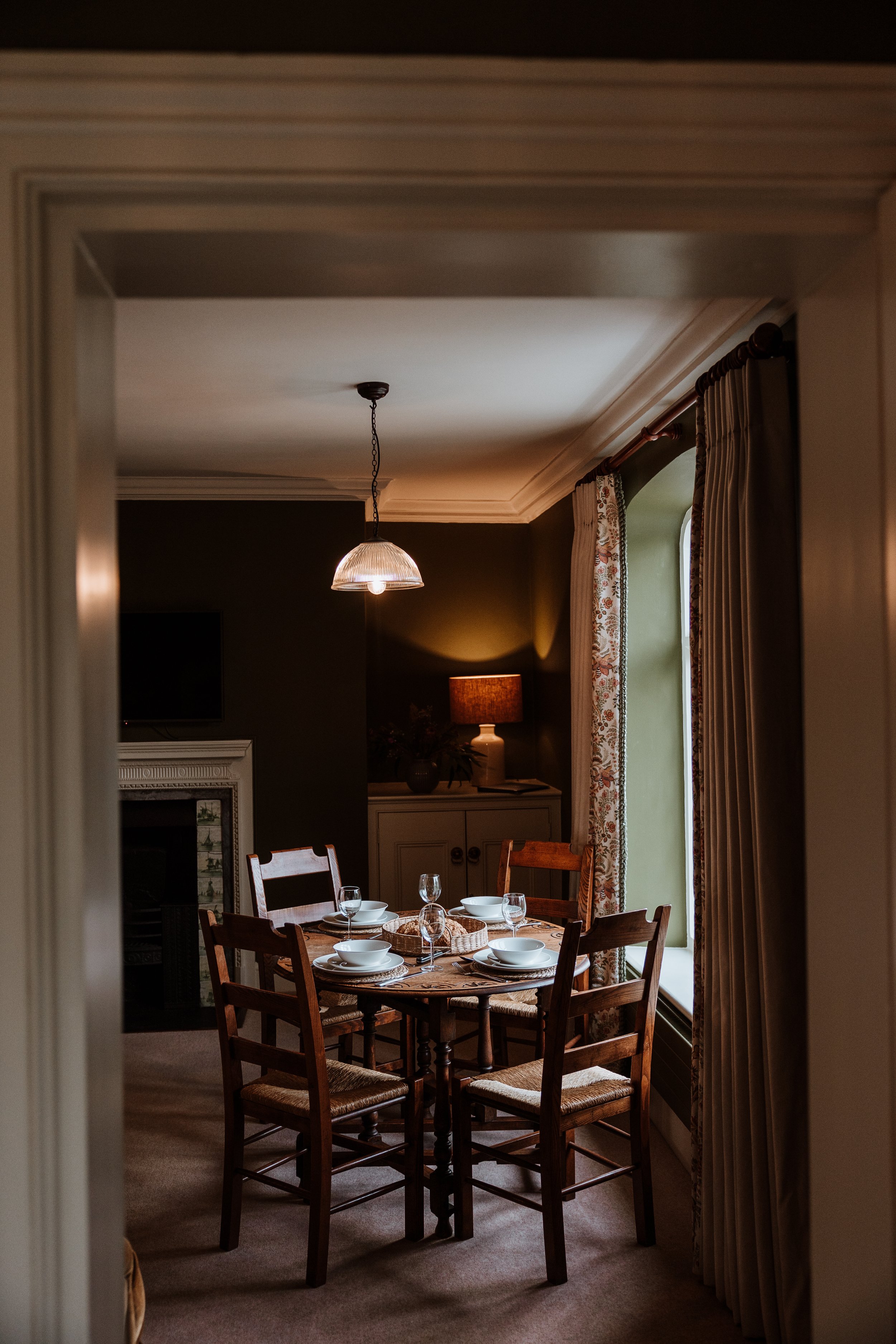 Dining room with wooden table set with white bowls and glasses, surrounded by wooden chairs, next to a window with drapes, warm lighting from a table lamp and hanging ceiling light. Interiors by Lucy Rebecca Photography.