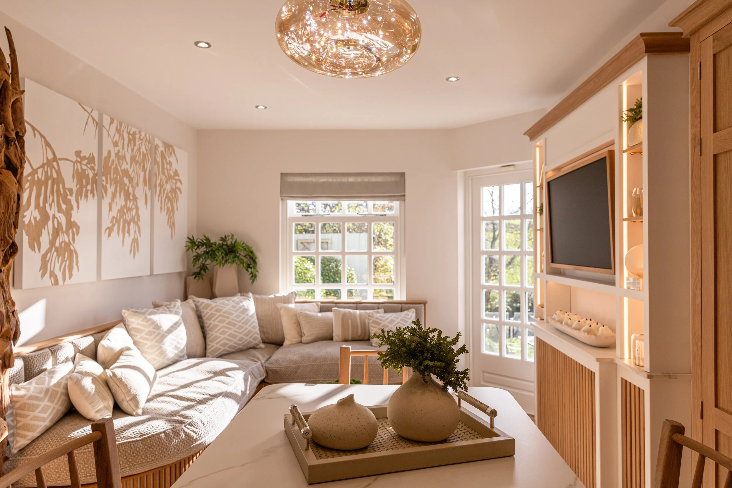 Bright living room with white walls, a large beige sectional sofa with patterned and plain pillows, a wooden coffee table with vases and greenery, a wooden entertainment center with a TV, decorative shelves, and warm lighting. Interior Photography.
