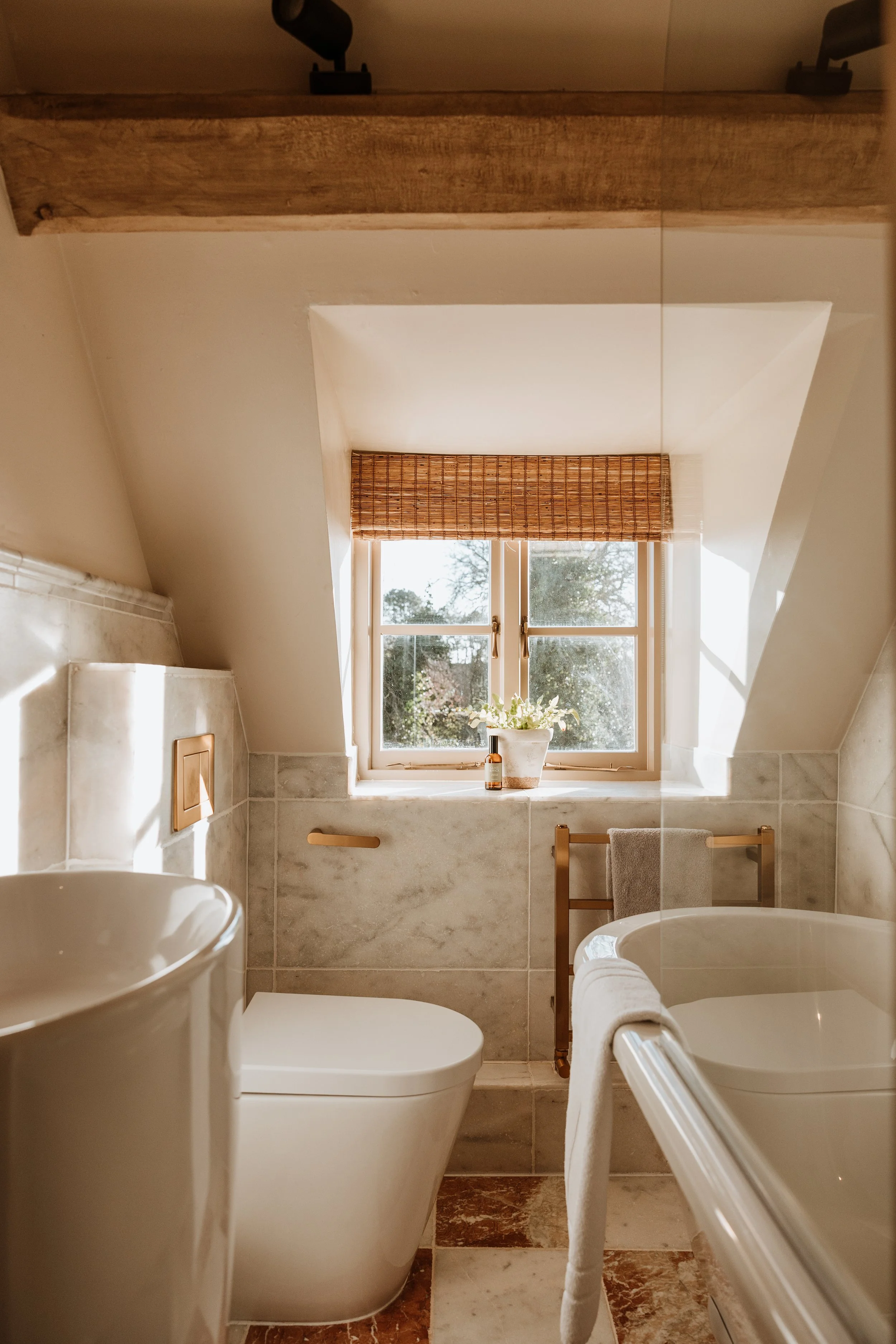 A small bathroom with a sloped ceiling, featuring a window with a bamboo shade, a potted plant, a bottle of soap, a modern toilet, and a bathtub with a towel hanging on a brass towel rack, all illuminated by natural light. Interiors by Lucy Rebecca 