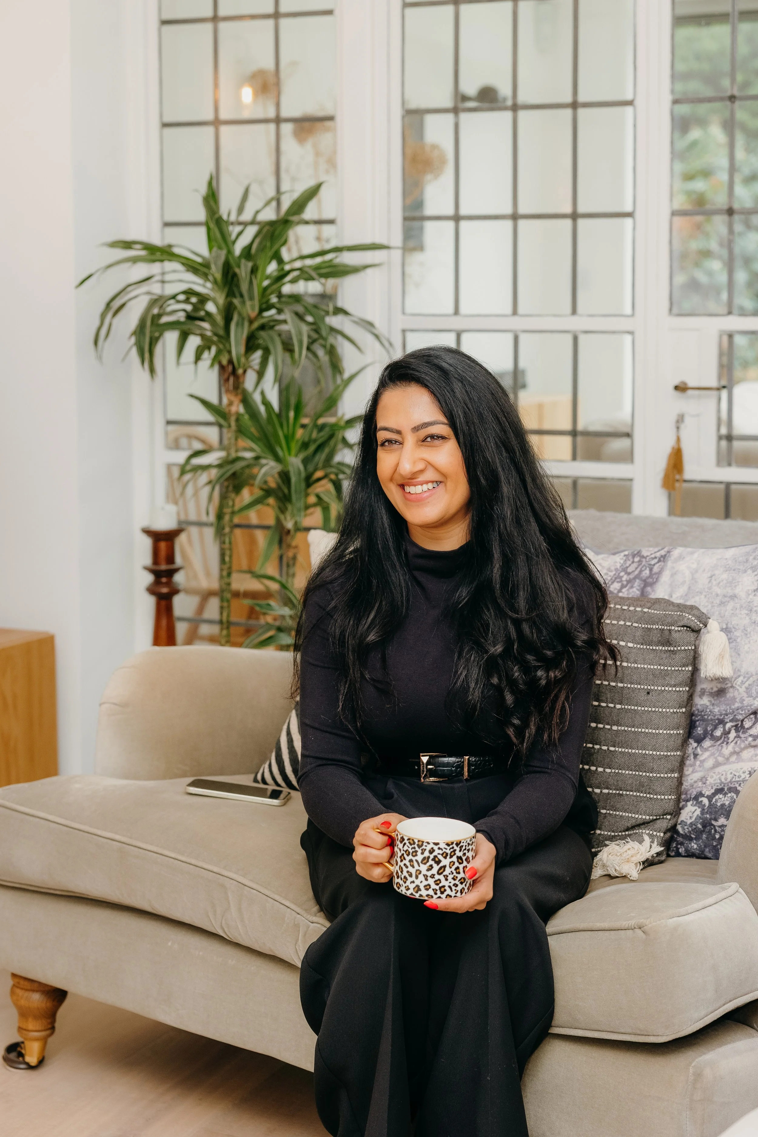 A woman with long black hair smiling and holding a mug with animal print sitting on a beige couch in a bright living room. Personal Branding by Lucy Rebecca Photography.