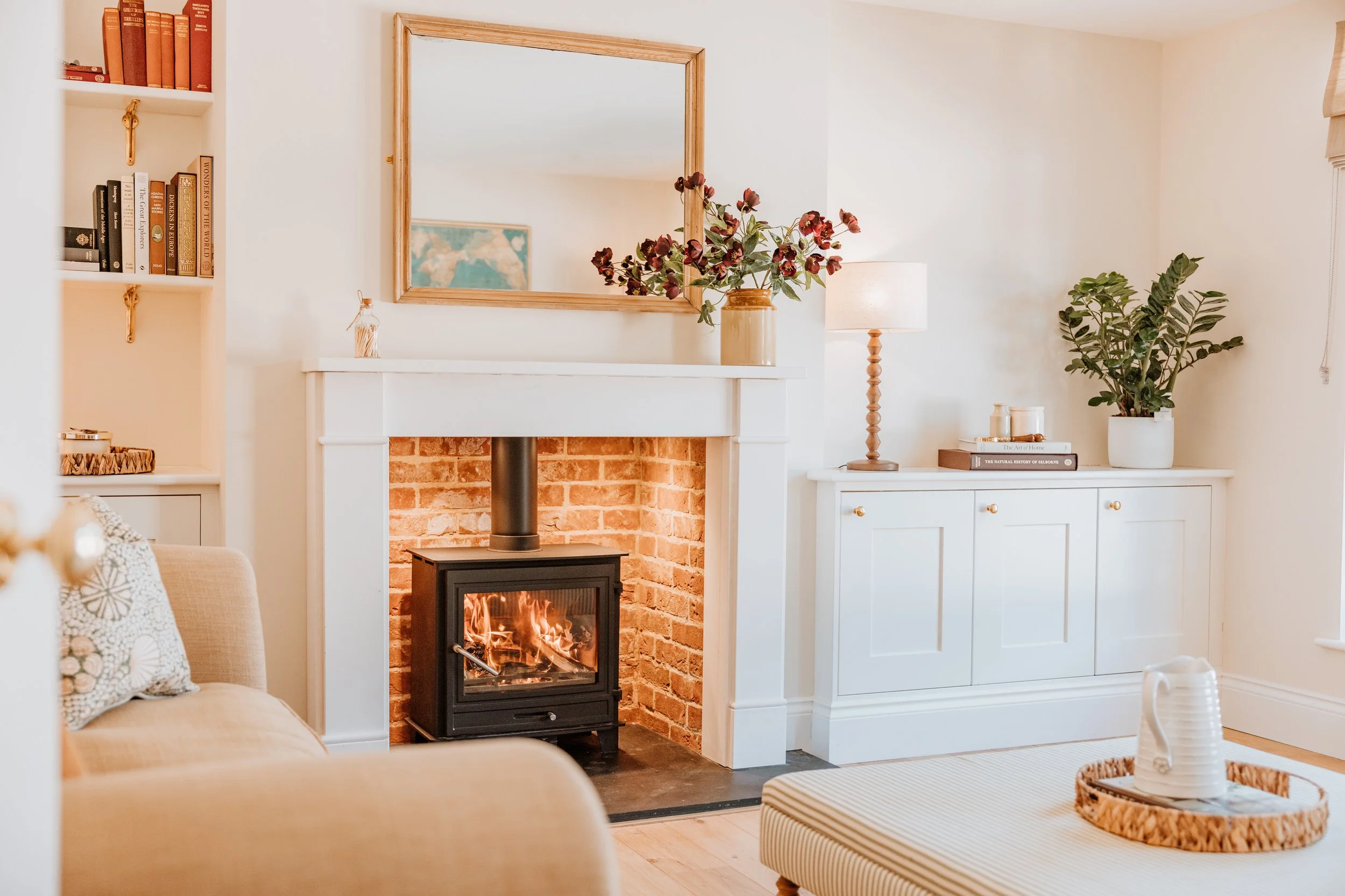 Living room with a cozy fireplace, beige sofa with patterned pillows, white cabinetry, potted plant, and decorative items, reflected in a large mirror. Interiors by Lucy Rebecca Photography.