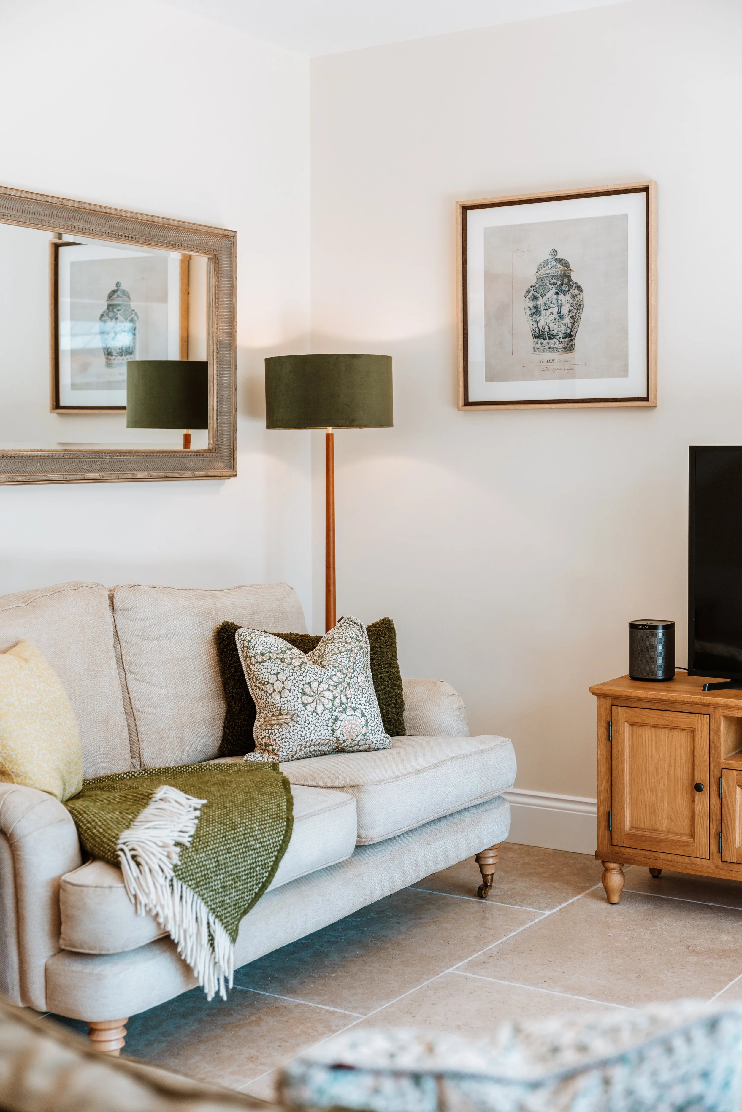 Living room with a beige sofa, green and patterned pillows, a wooden TV stand with a black TV and smart speaker, a floor lamp with a green shade, a large mirror, and framed art on the wall. Interiors by Lucy Rebecca Photography.