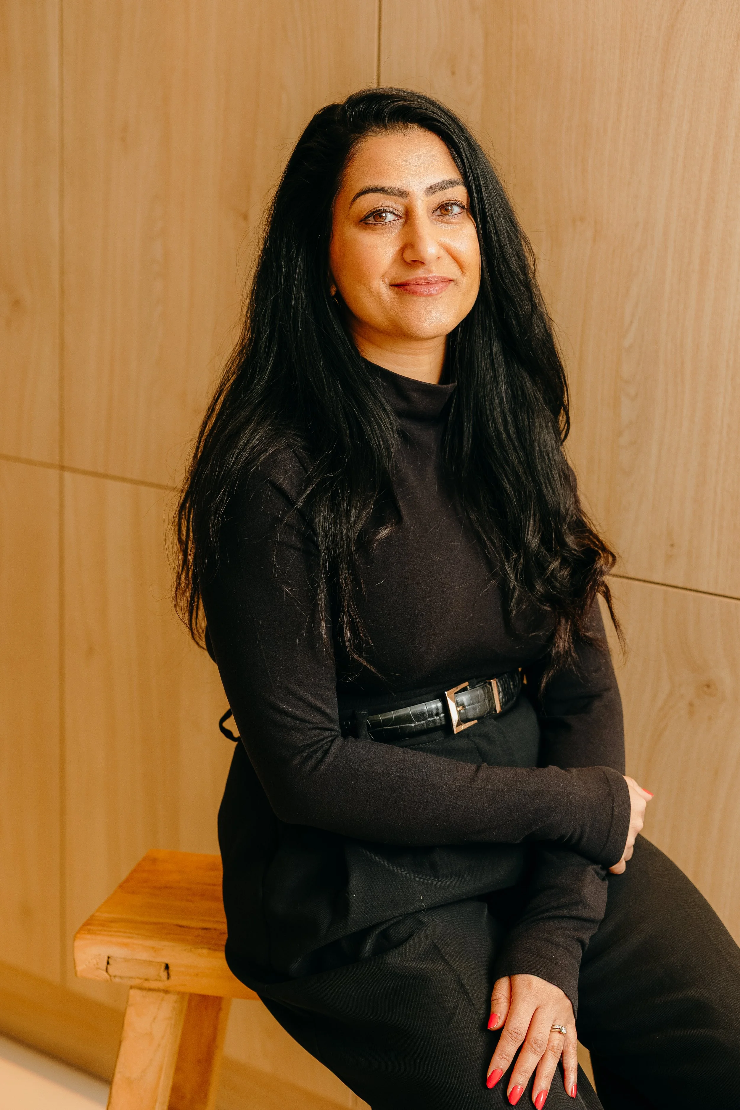 A woman with long black hair, wearing a black turtleneck and black pants, sitting on a wooden stool against a wooden wall, smiling at the camera. Personal Branding by Lucy Rebecca Photography.