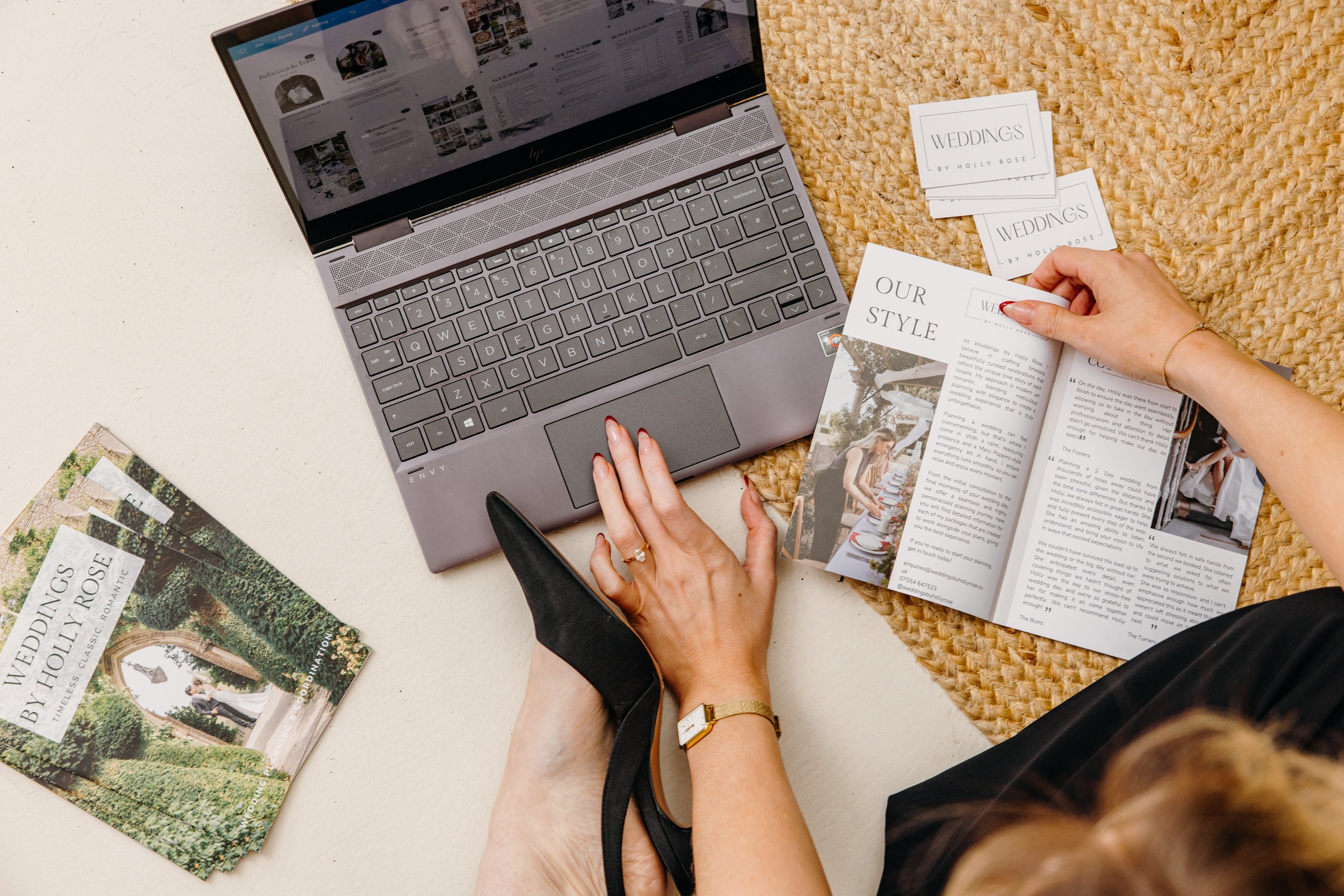A person preparing wedding brochures and printed materials on a woven mat, with an open laptop displaying a website, and additional brochures and a magazine on the floor.Personal Branding by Lucy Rebecca Photography.