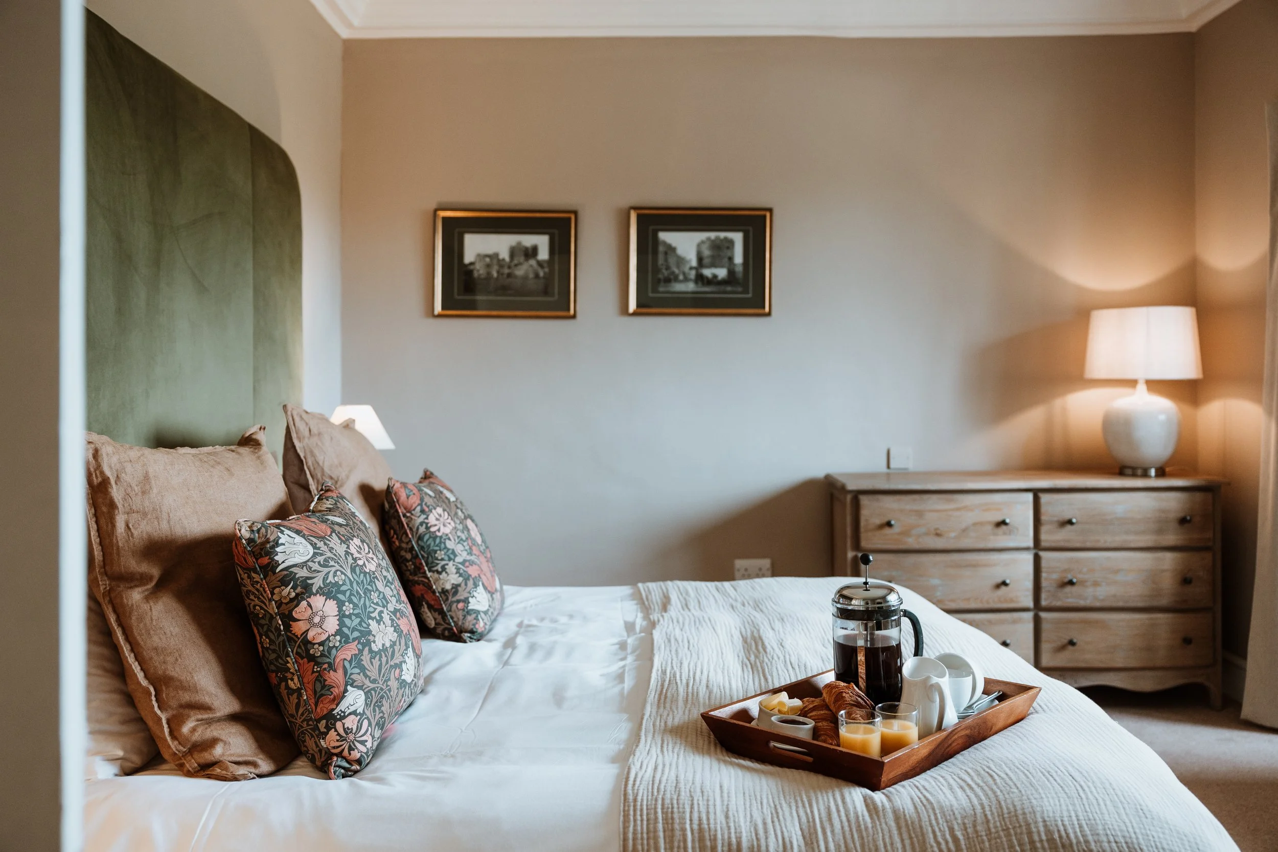 A cozy bedroom with a bed that has patterned pillows, a tray with breakfast items, and a wooden dresser with a lamp, two framed pictures on the wall. Interiors by Lucy Rebecca Photography.