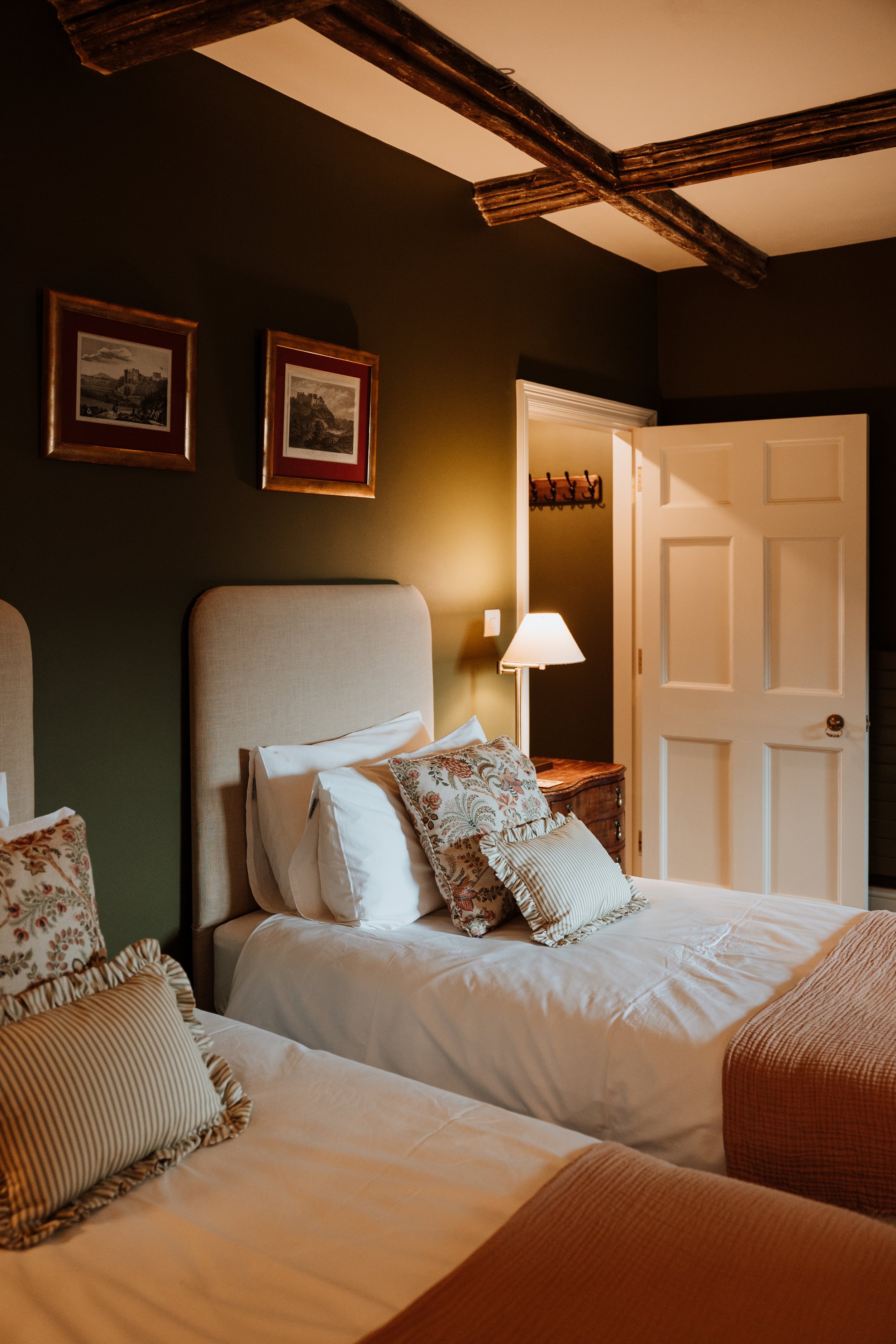 A cozy bedroom with two beds, beige upholstered headboards, floral and striped pillows, a nightstand with a lamp, framed artwork on a dark green wall, and an open door leading to a small closet with a coat rack. Interiors by Lucy Rebecca Photography.