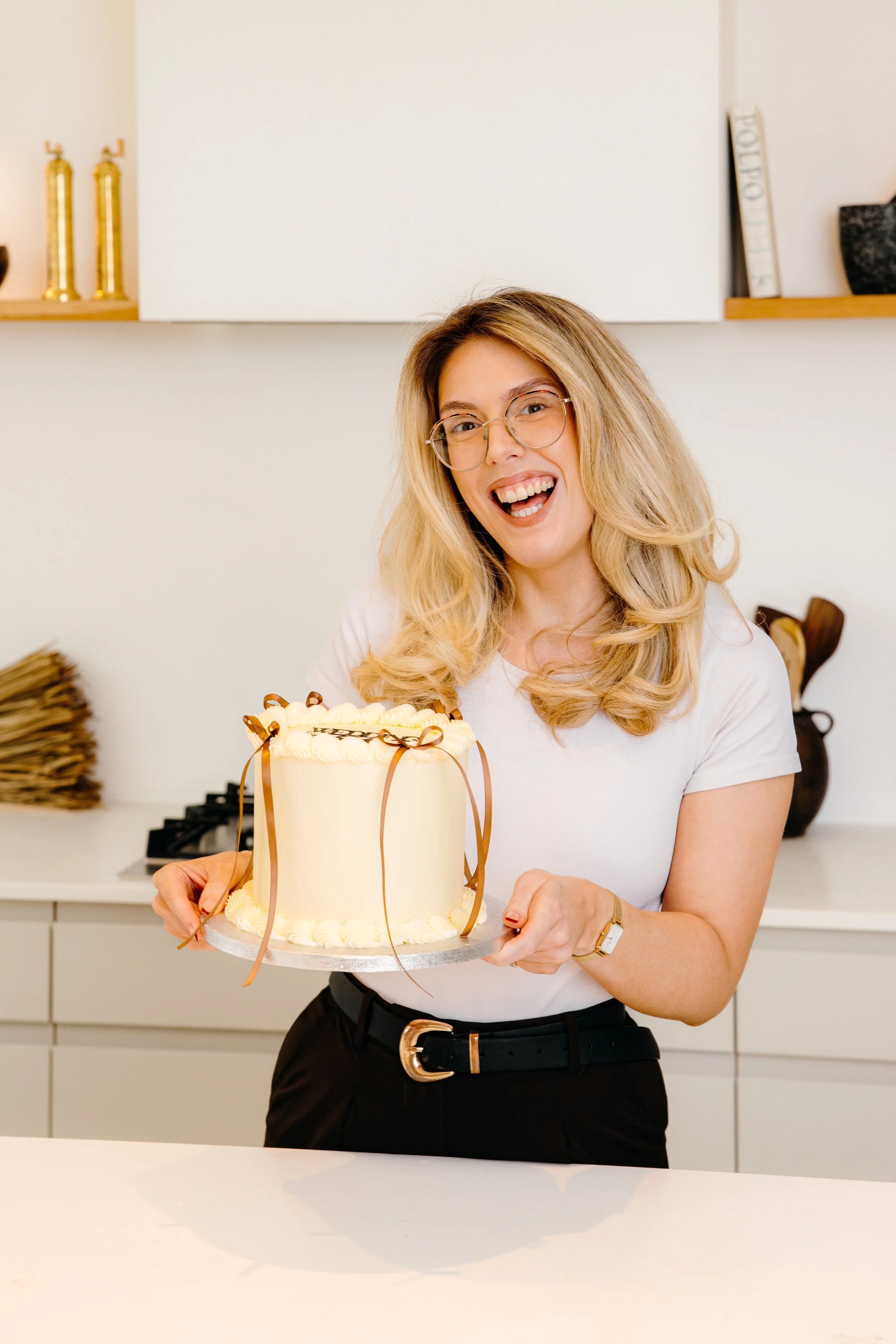 A woman with blonde hair, glasses, and a white shirt holding a lemon cake decorated with chocolate ribbons in a modern kitchen. Personal Branding by Lucy Rebecca Photography.