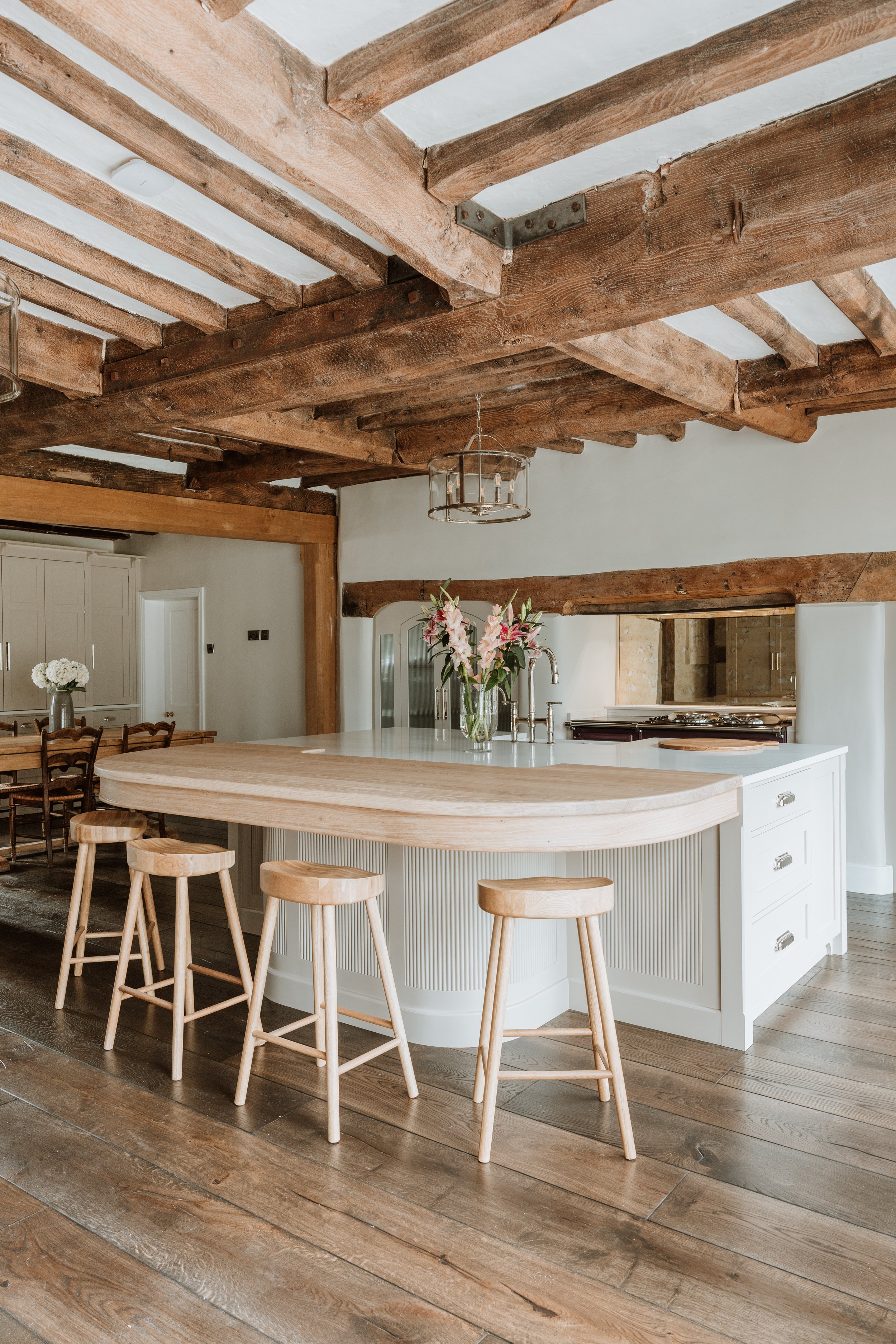 A modern kitchen and dining area with a wooden ceiling and beams, a white kitchen island with a flower vase, four wooden bar stools, a chandelier, and a wooden dining table with chairs. Interiors by Lucy Rebecca Photography.