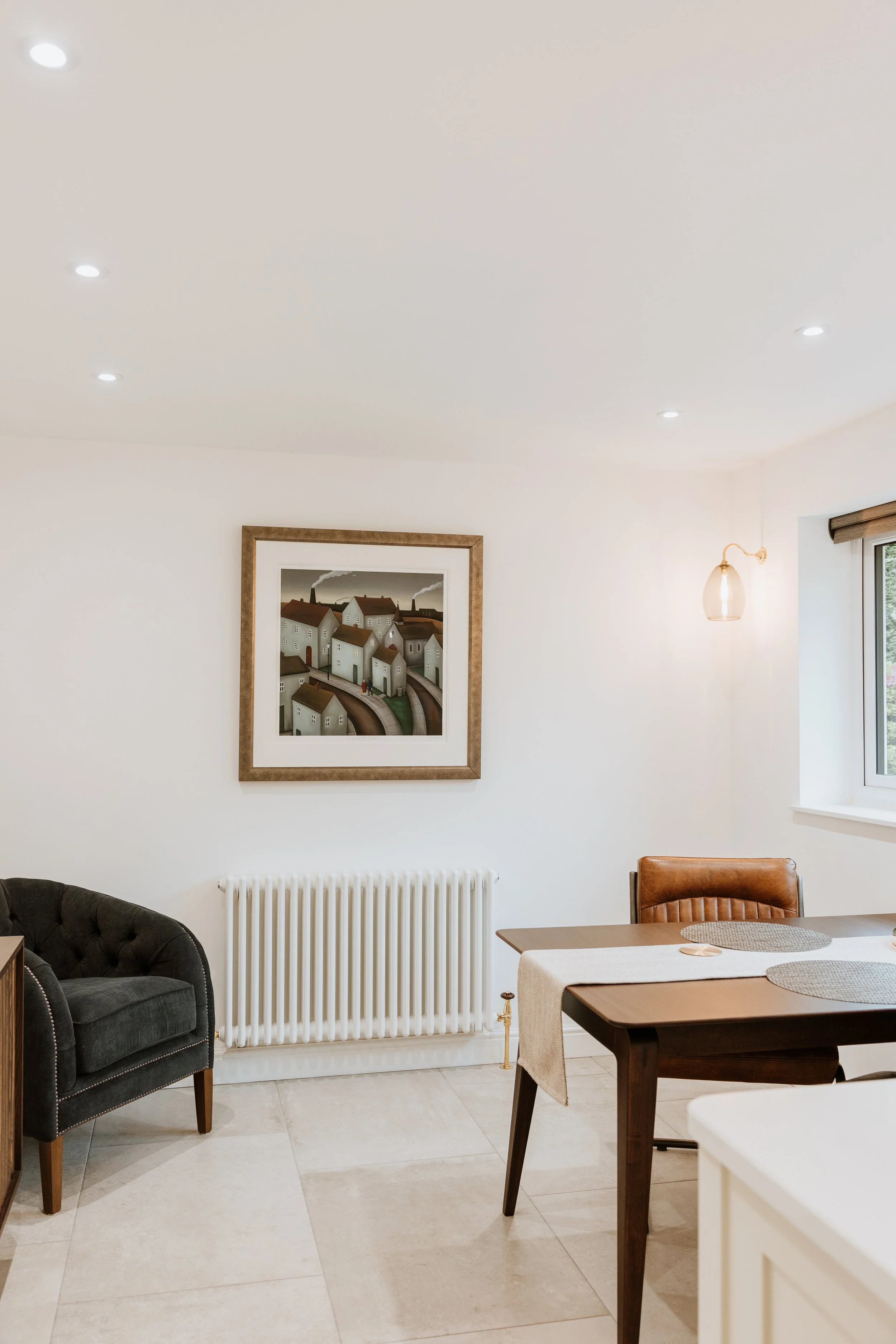 A dining area with a painting of a town on the wall, a window with a blinds, a brown leather chair, a black upholstered armchair, a radiator, a dining table with a beige runner, and round placemats. Interiors by Lucy Rebecca Photography.