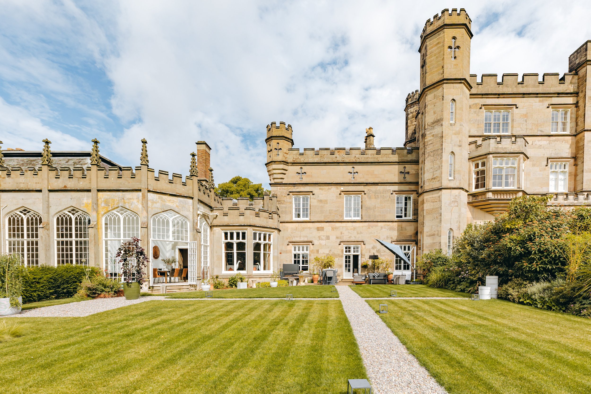 A large stone castle with turrets and towers, surrounded by a well-maintained lawn and garden, under a partly cloudy sky. Interiors by Lucy Rebecca Photography.