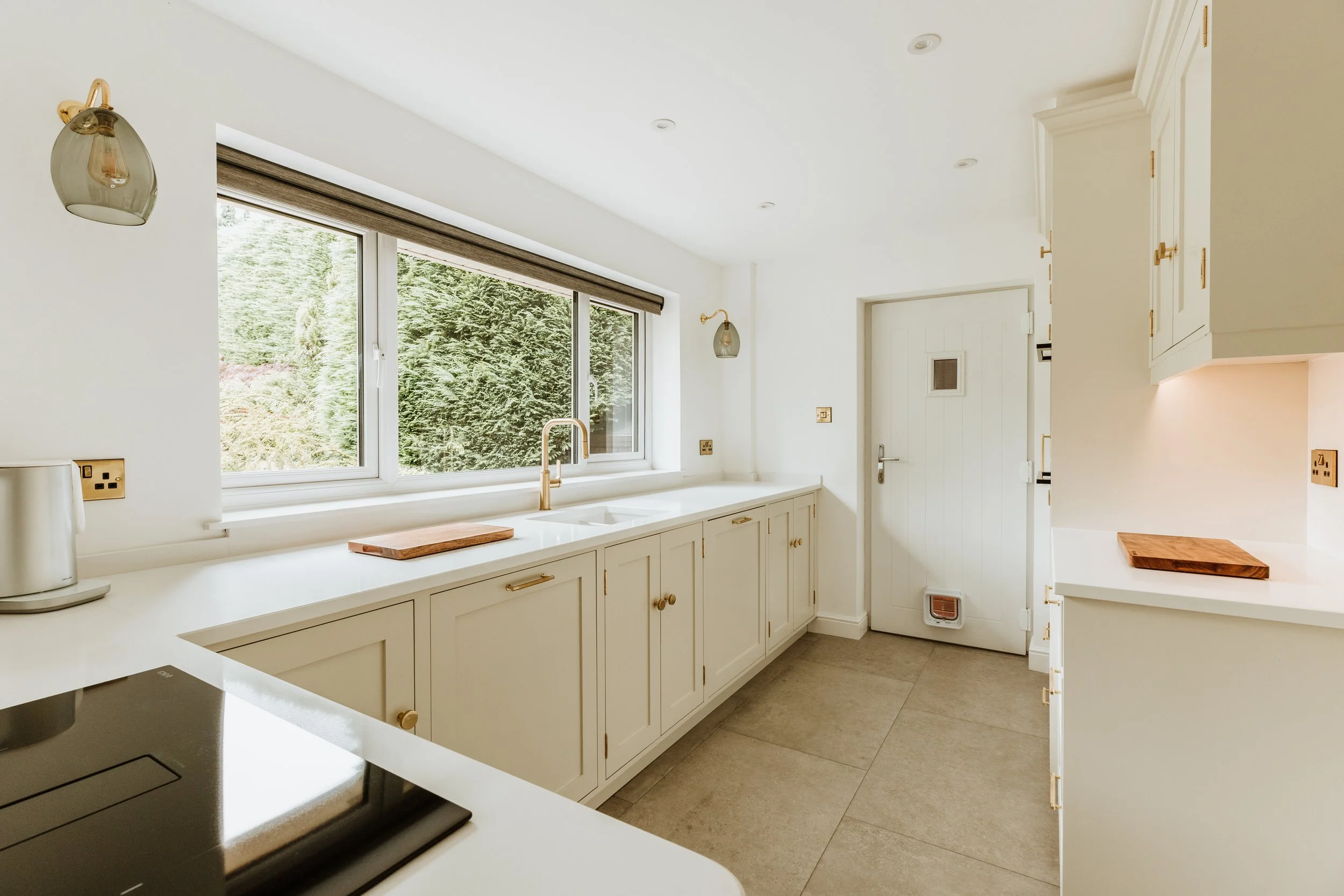Bright kitchen with white cabinets, large window overlooking greenery, beige countertops, and a stovetop. Interiors by Lucy Rebecca Photography.
