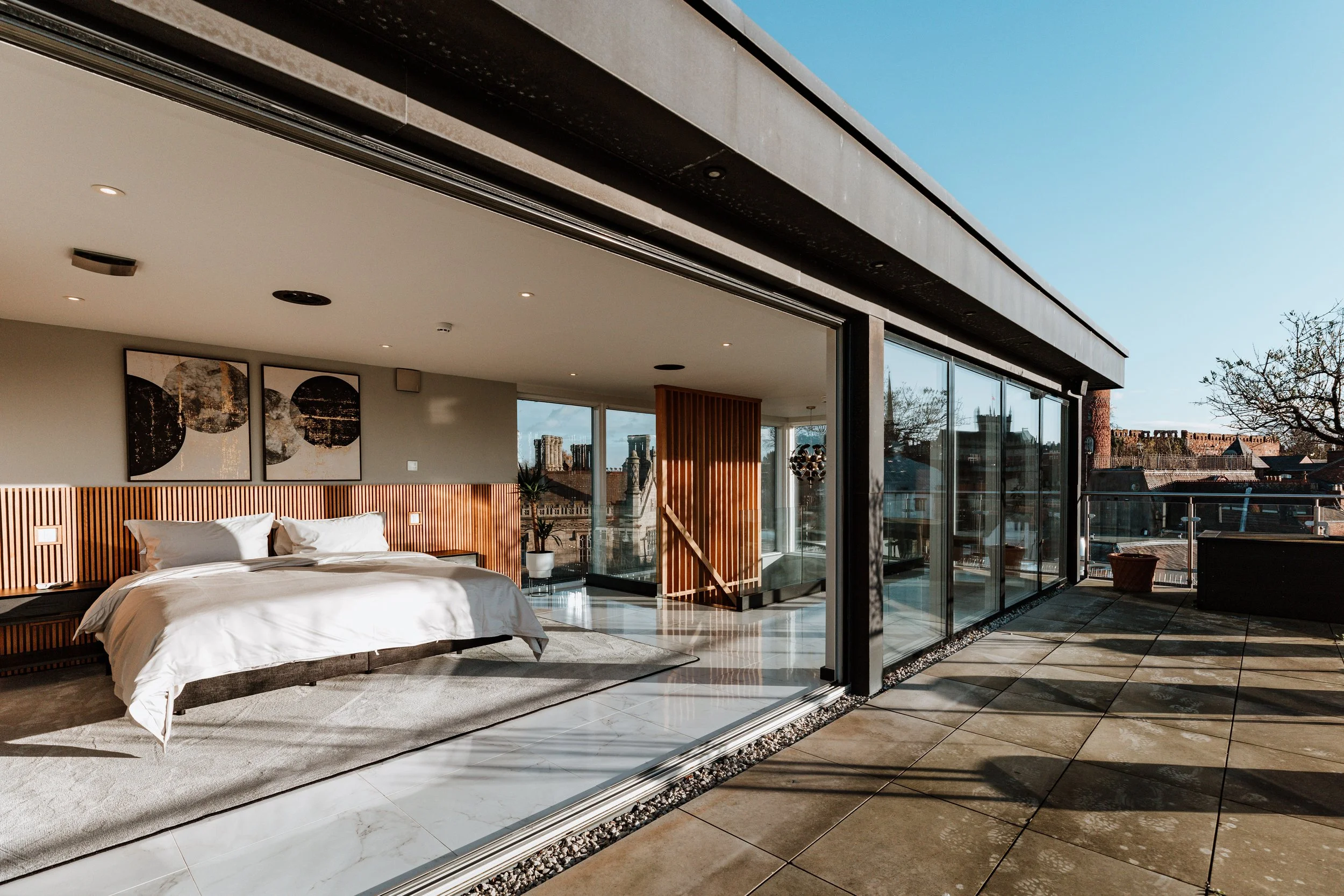 Modern bedroom with large sliding glass doors opening to a balcony, featuring a bed with white linens, abstract artwork, and hardwood accents.  Interiors by Lucy Rebecca Photography.