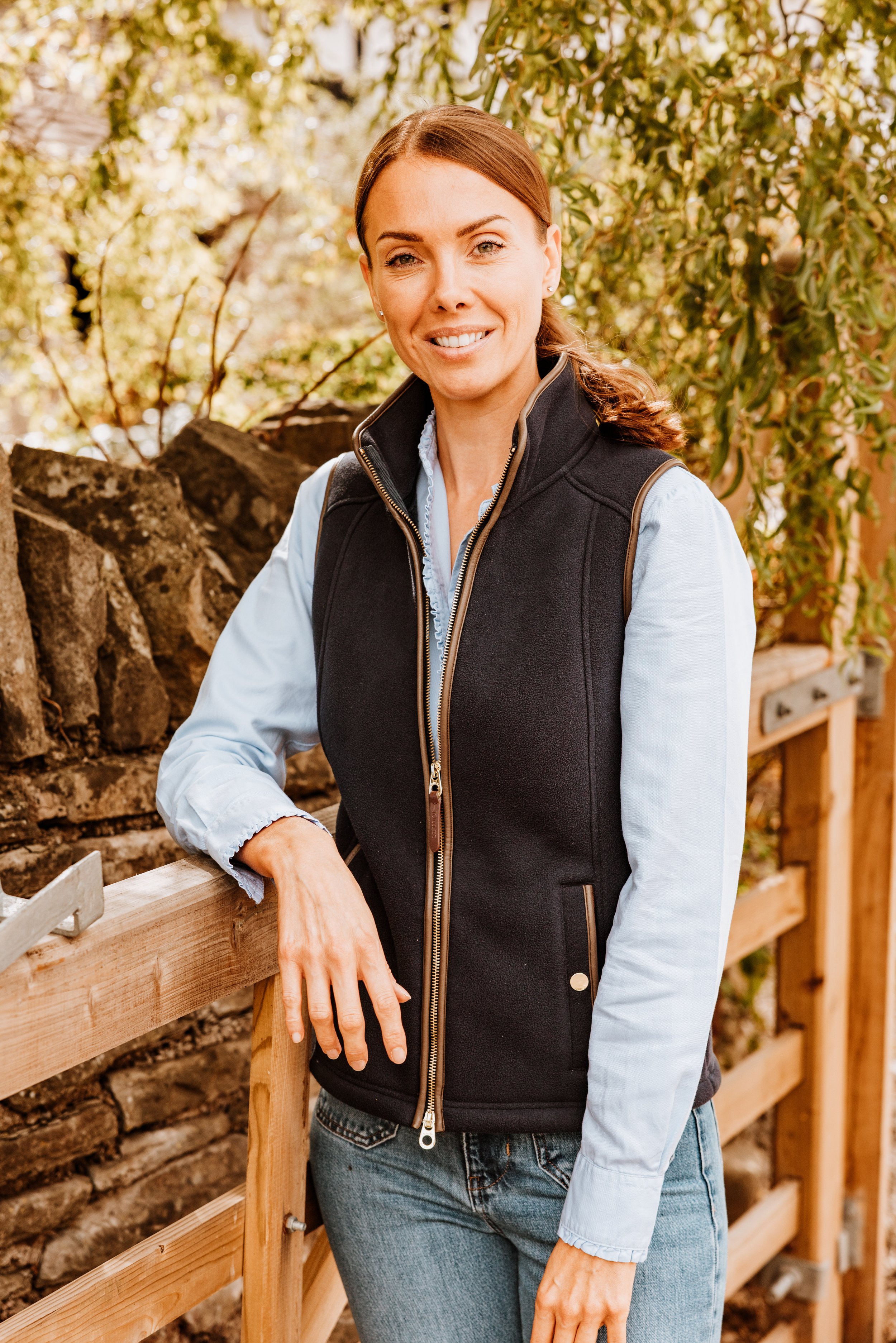 A woman outdoors near a wooden fence, wearing a black vest over a light blue shirt, with brown hair and a friendly smile. Personal Branding by Lucy Rebecca Photography.